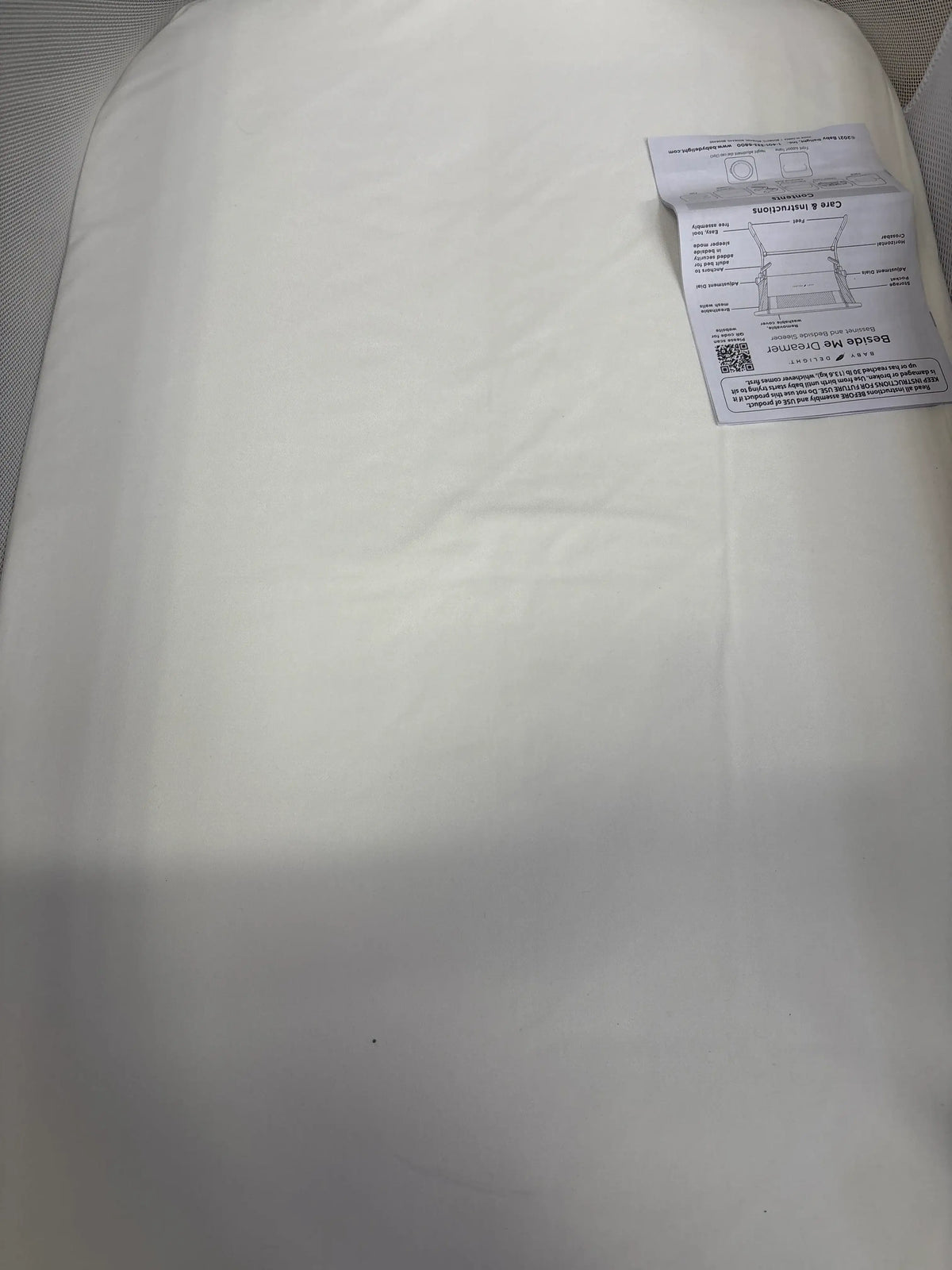 Top-down view of Baby Delight Beside Me Dreamer bedside bassinet showcasing mattress with white fitted sheet and instruction manual in corner