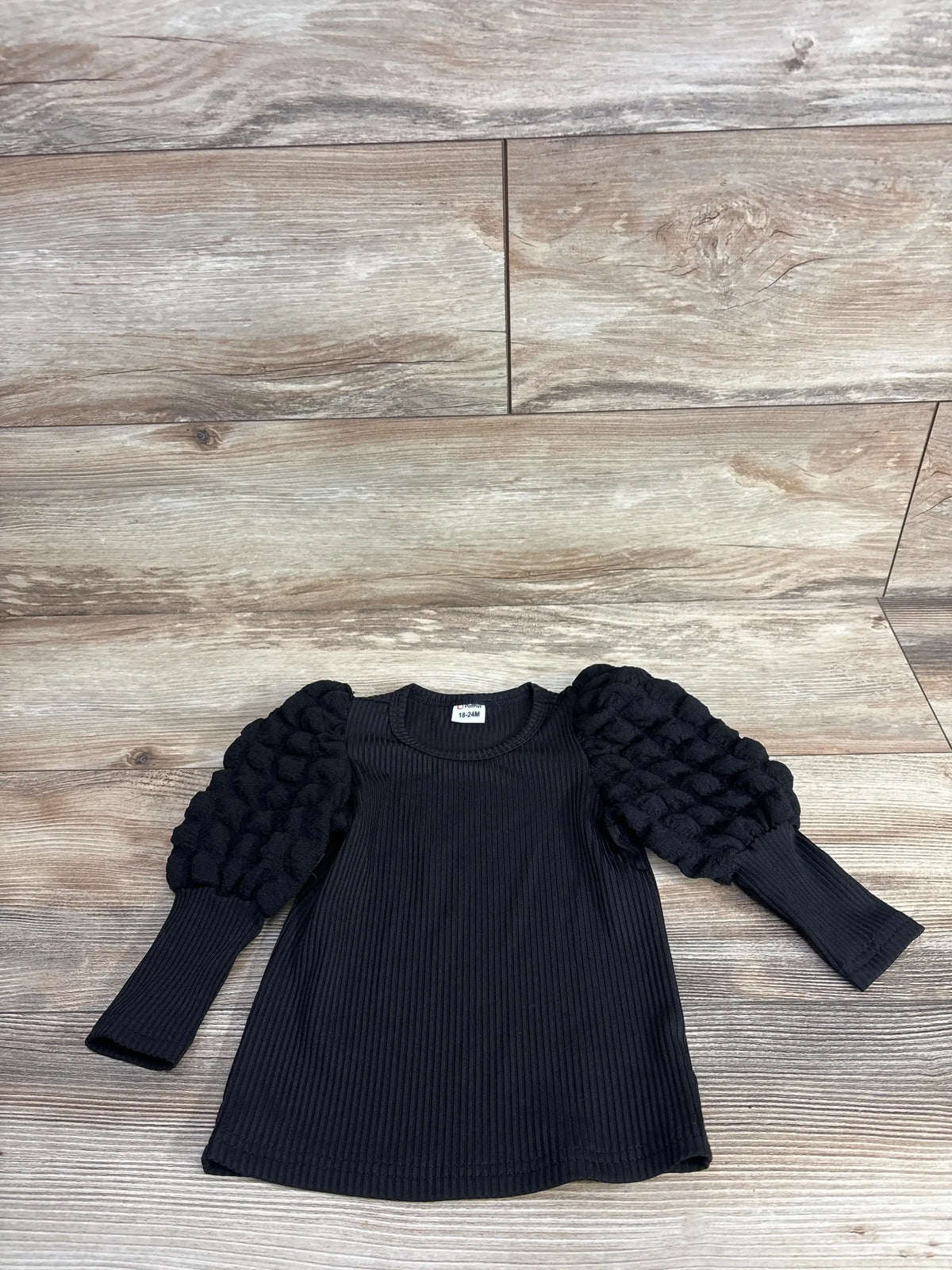 Front view of Pat Pat black ribbed puff sleeve shirt, size 18 to 24 months, showcasing textured long sleeves on wood background