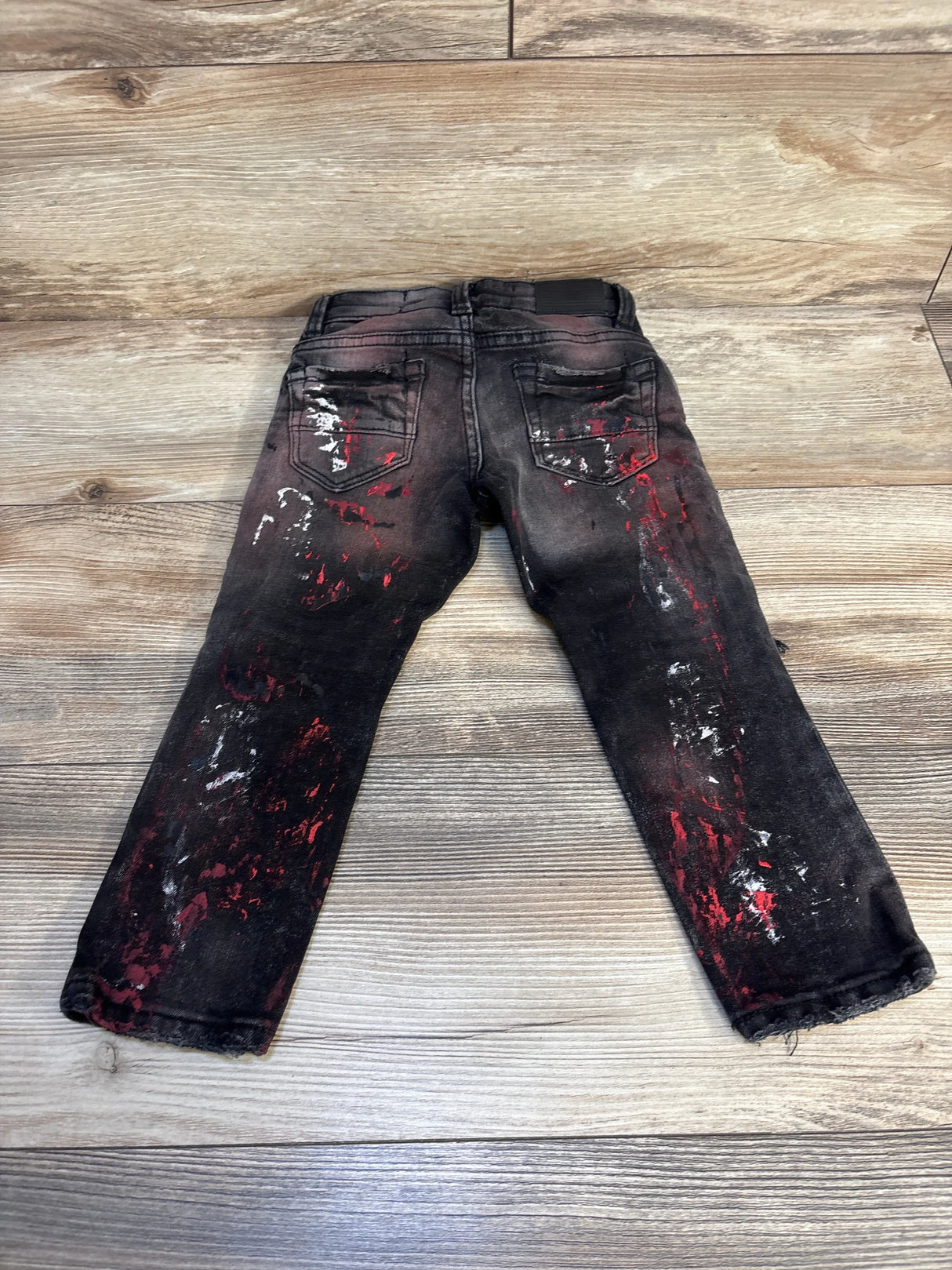 Black kids jeans with red and white paint splatter, back pockets, displayed on wood floor