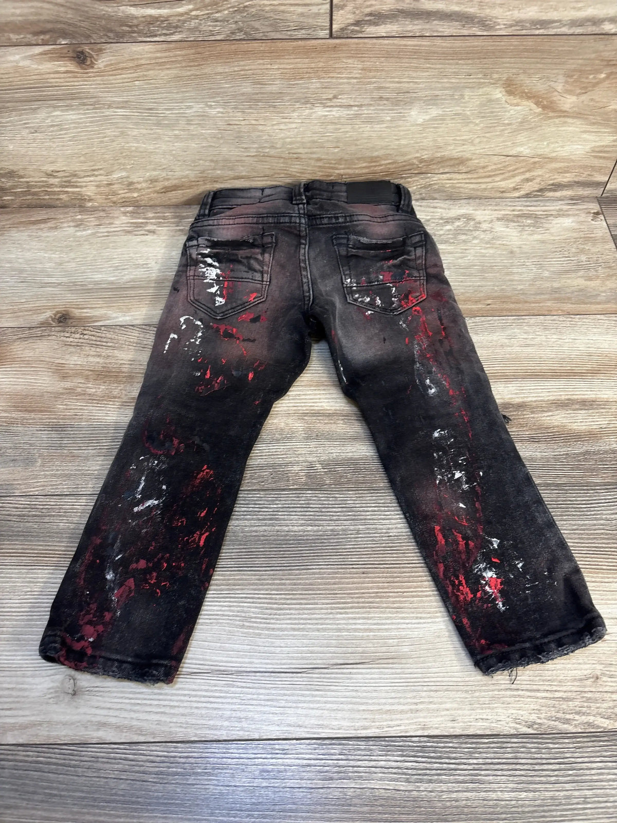 Black kids jeans with red and white paint splatter, back pockets, displayed on wood floor