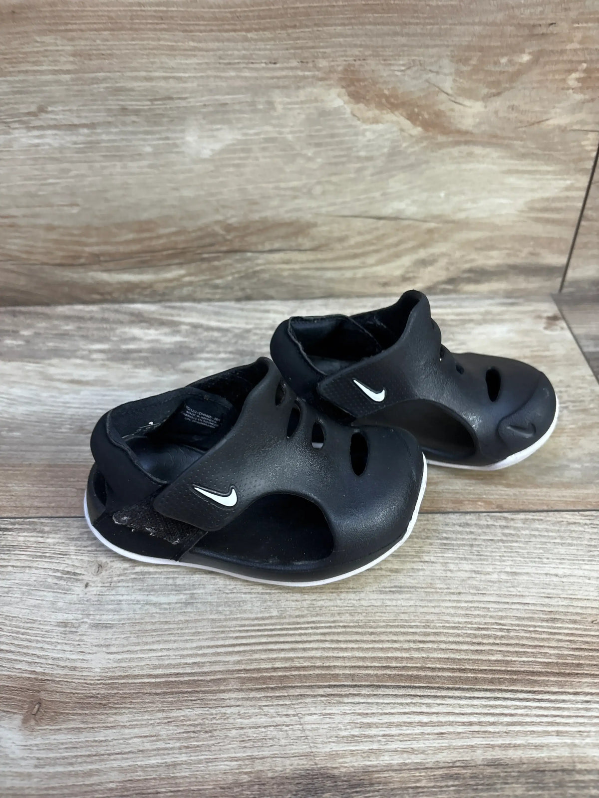 Three-quarter view of Nike Sunray Protect 3 TD toddler sandals in black with white swoosh, velcro strap and cutout vents on wood background