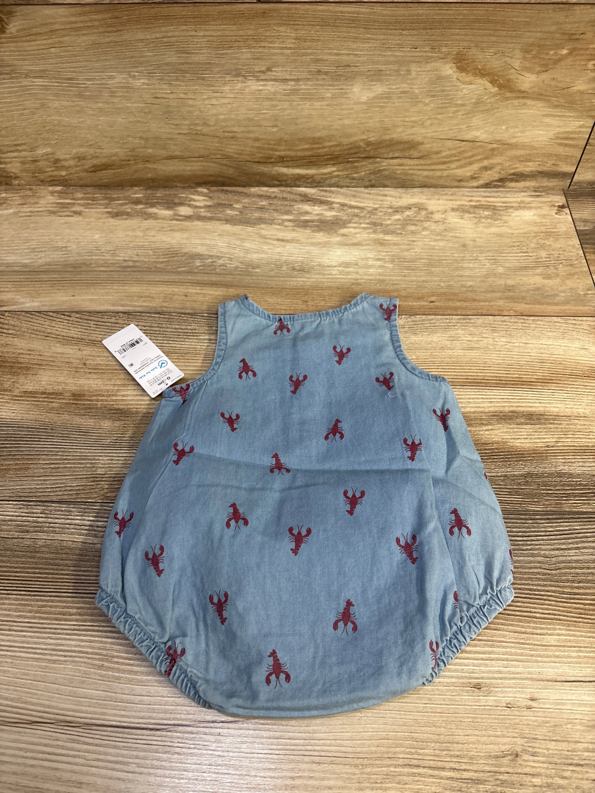 Flat lay of blue chambray bubble romper with red lobster print, sleeveless with elastic leg openings on wood background