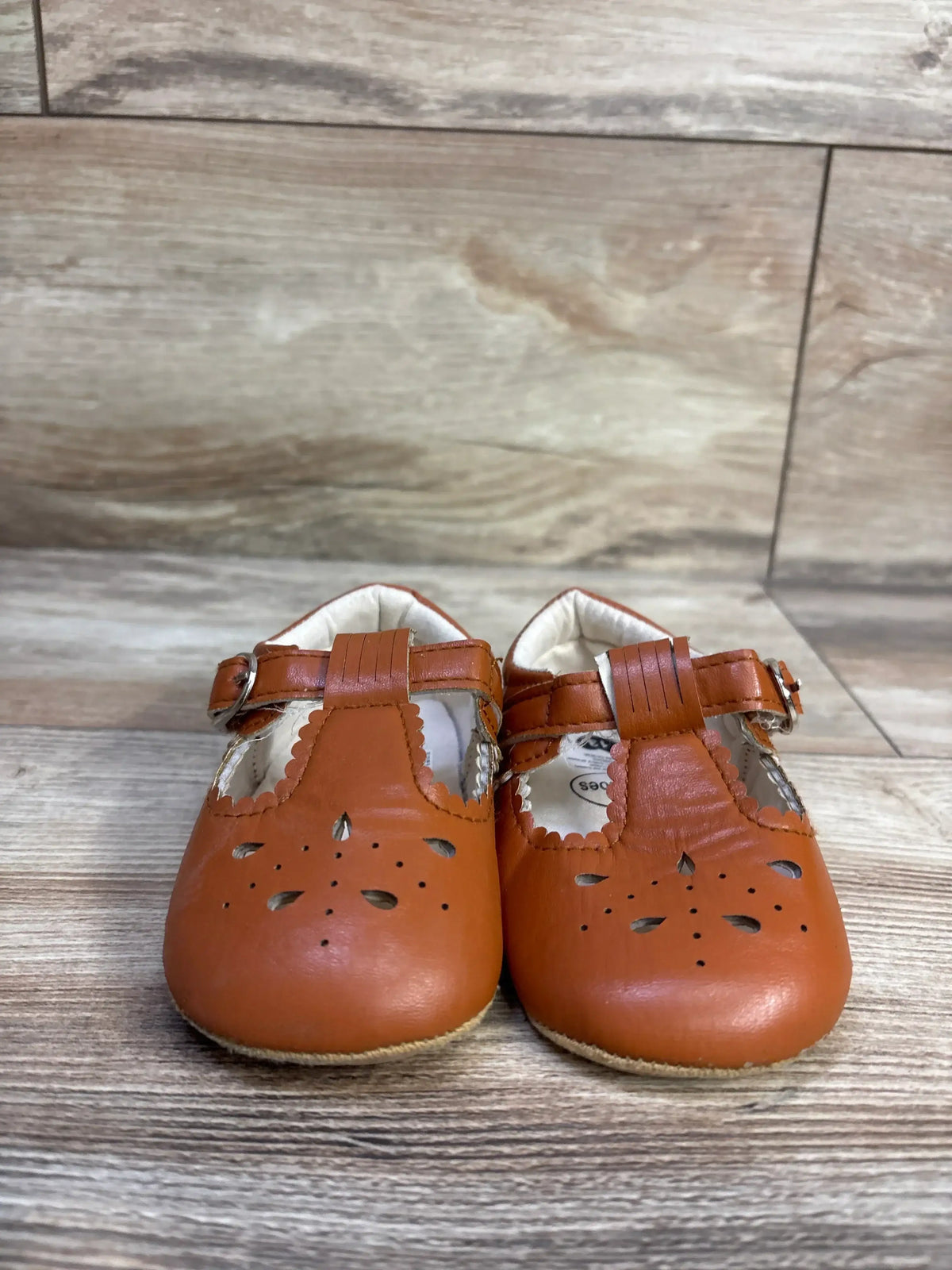 Front view of Sienna baby sandals in chestnut, T-strap with perforated floral cutouts on light wood background