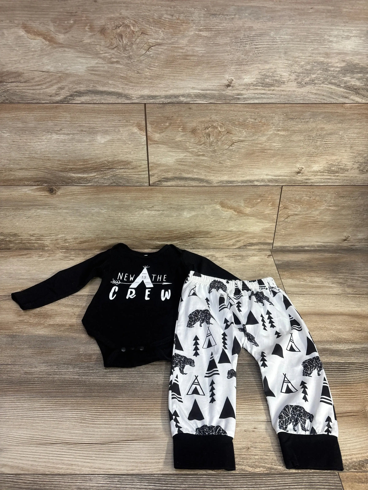Flat lay of black long-sleeve bodysuit reading NEW TO THE CREW with white pants printed with black bears and teepees on wood floor