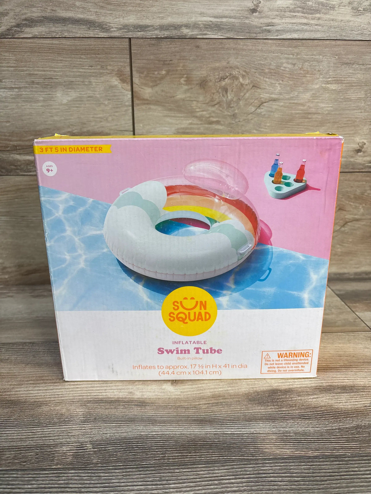 Front view of Sun Squad inflatable rainbow swim tube box showing rainbow tube graphic, built-in pillow and 41 in diameter label on wood floor