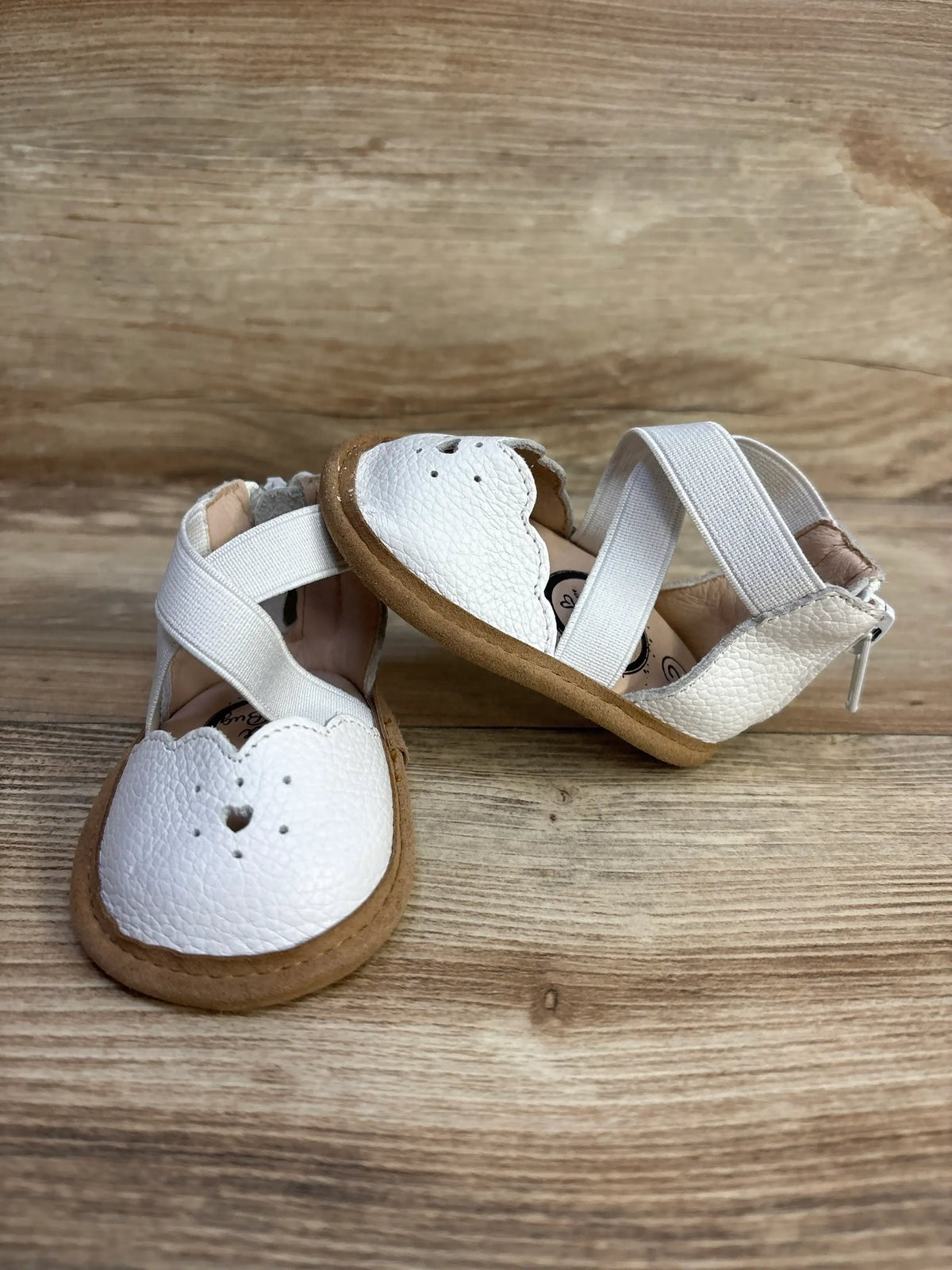 Three-quarter view of Little Love Bug The Sophia Shoes size 2c in white with scalloped heart cutout and elastic crossover straps on a wood plank background