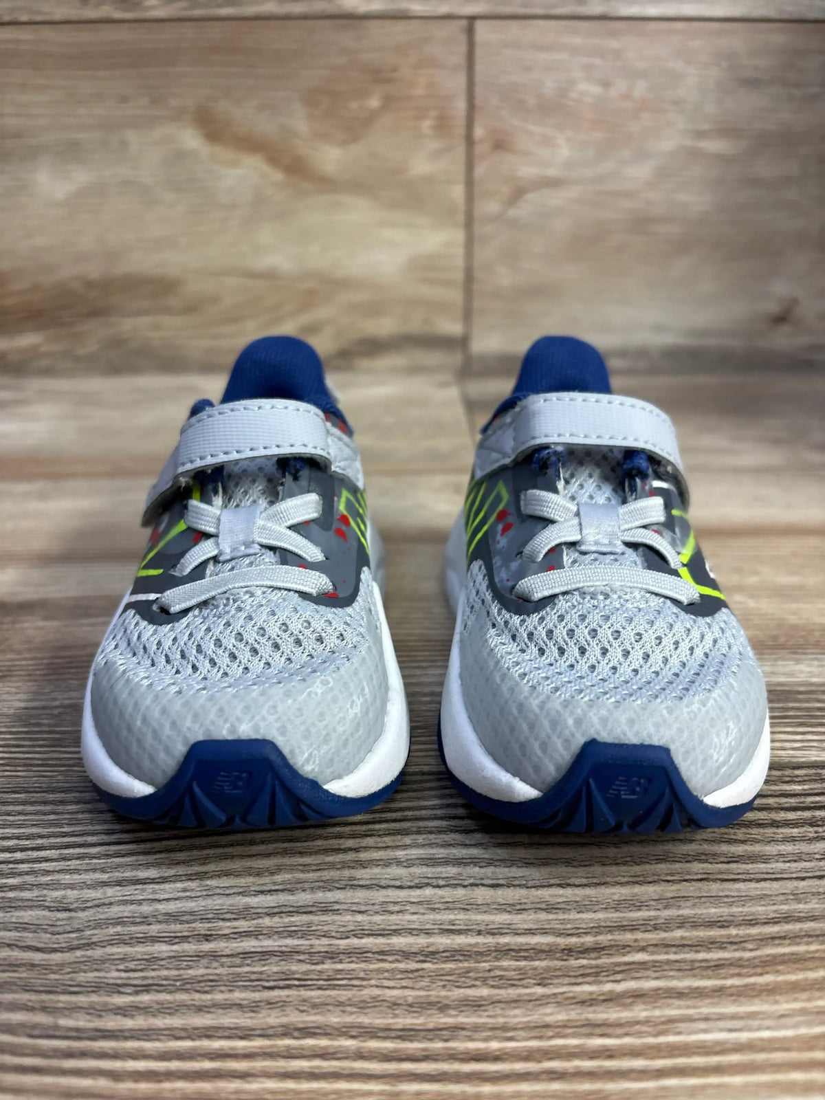 Front view of New Balance Rave Run v2 kids’ sneakers showing gray mesh upper, Velcro strap, and blue accents on wood floor
