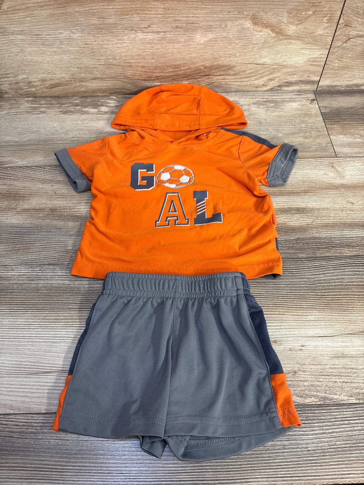 Flat lay of orange hooded toddler shirt size 12 months with GOAL soccer graphic and gray elastic-waist shorts on wood floor