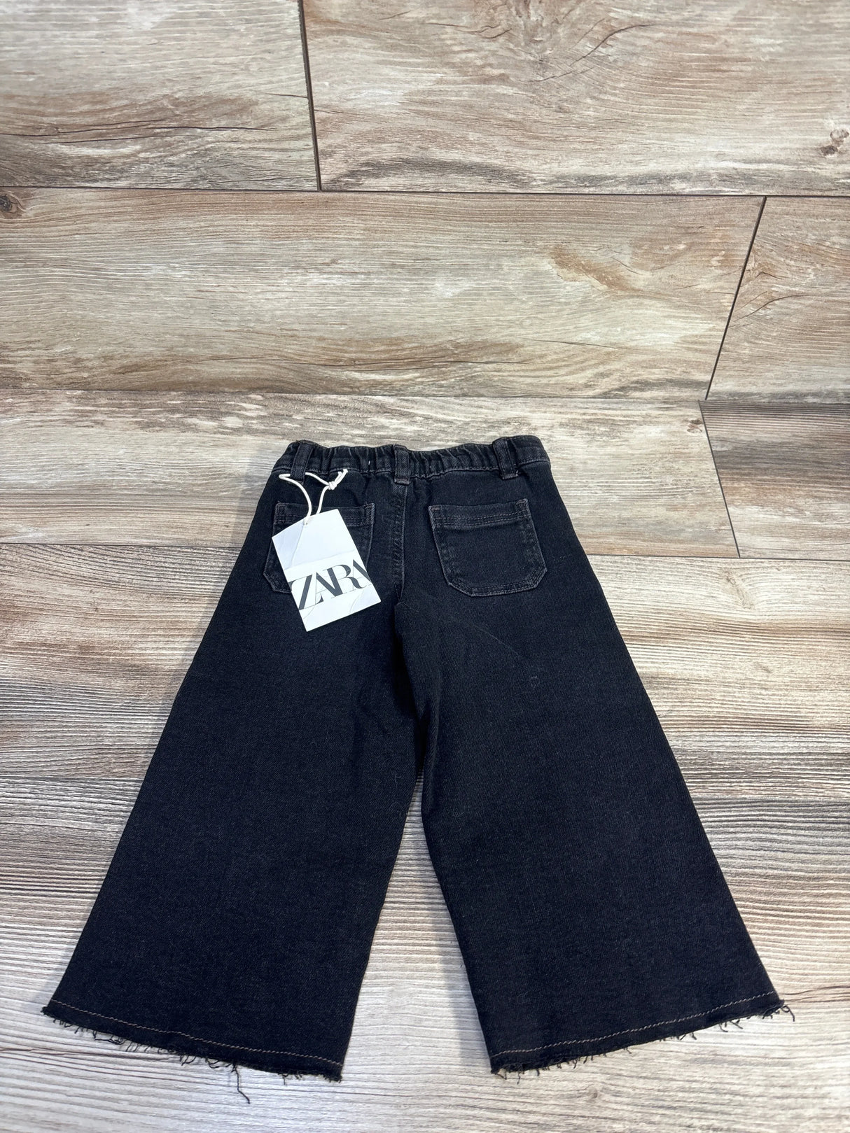 Back view of black Zara toddler wide-leg jeans (18–24m) showing frayed hems, elastic waistband and Zara tag on wood floor