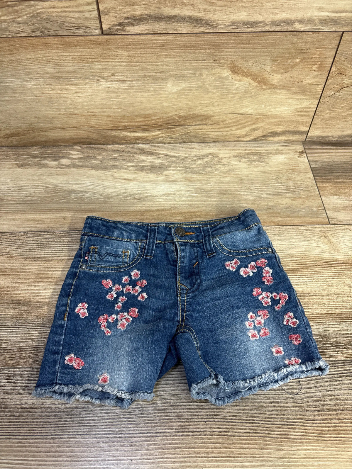 Front view of Vigoss Malibu mid-rise shorts showcasing pink floral embroidery and frayed hems on a wood-look floor