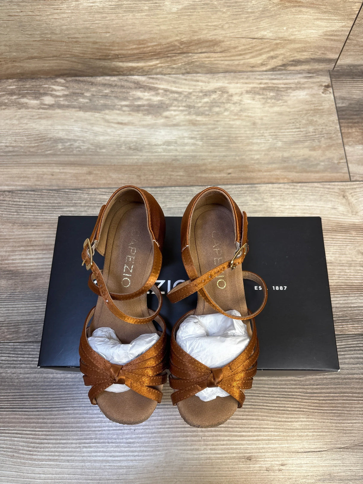 Brown Capezio child dance sandals with ankle straps on wood floor, displayed on black box