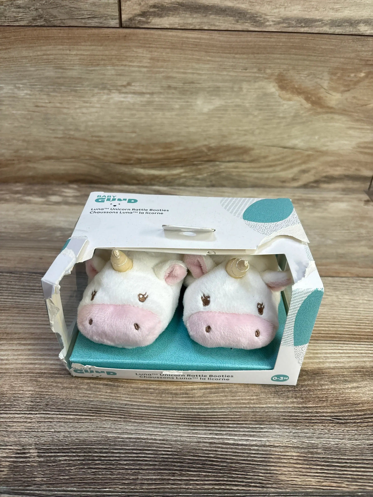 Front view of GUND Luna unicorn plush rattle booties showing unicorn faces and gold horns in box on wood surface