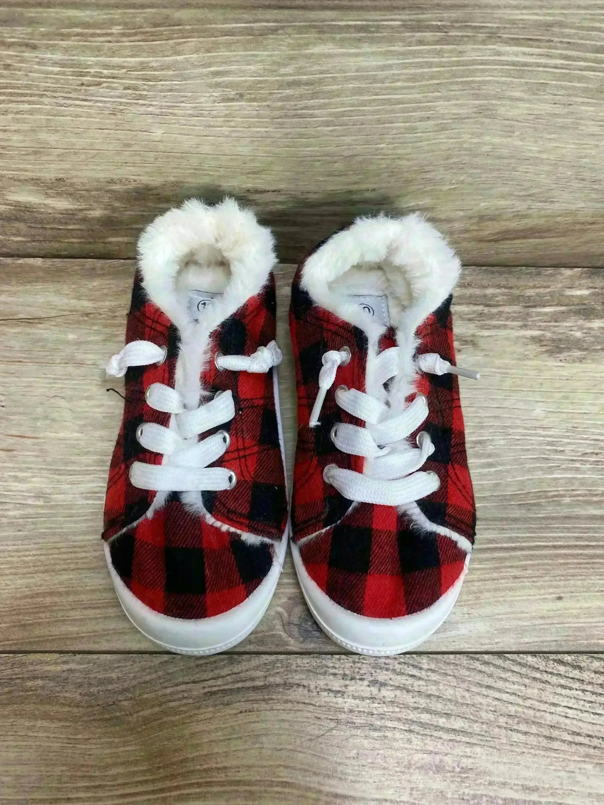 Front view of toddler Bumble + Birdie slip-on sneakers showing red and black buffalo plaid, white faux-fur lining and white laces on wood floor