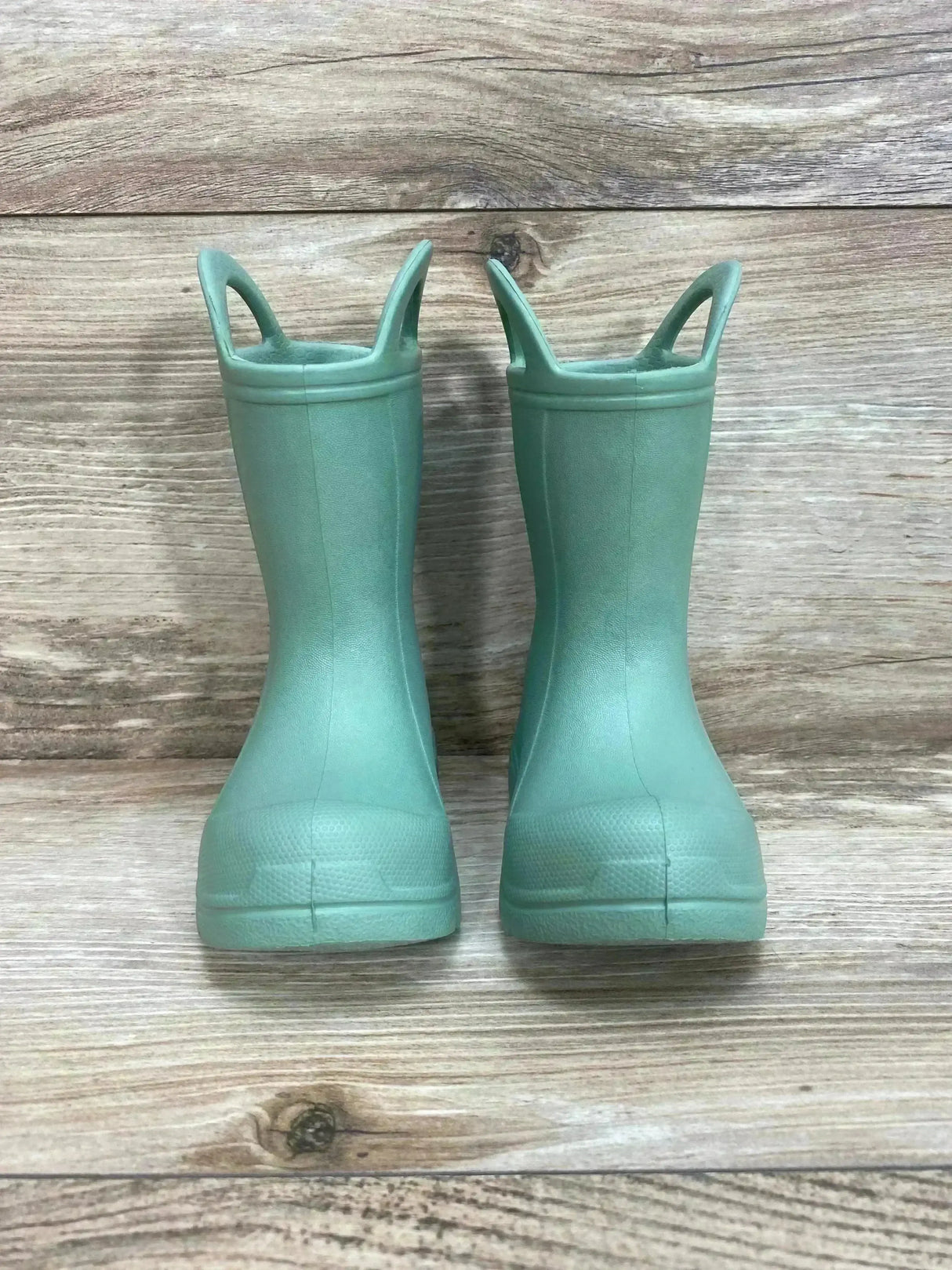 Front view of BEARPAW toddler green EVA rain boots showcasing easy pull-on handles on light wood background