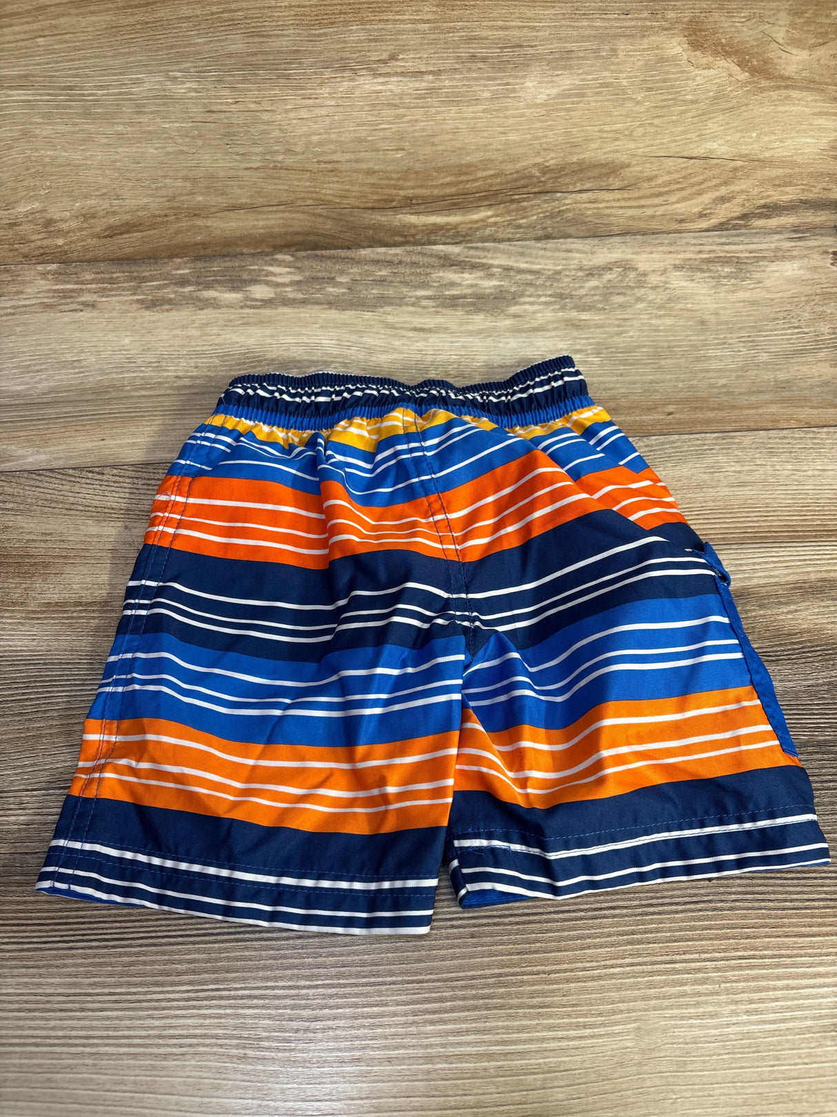Flat lay of UV Skinz Striped Swim Trunks in navy sz 3T showcasing blue orange and white stripes with elastic waistband on wood plank background