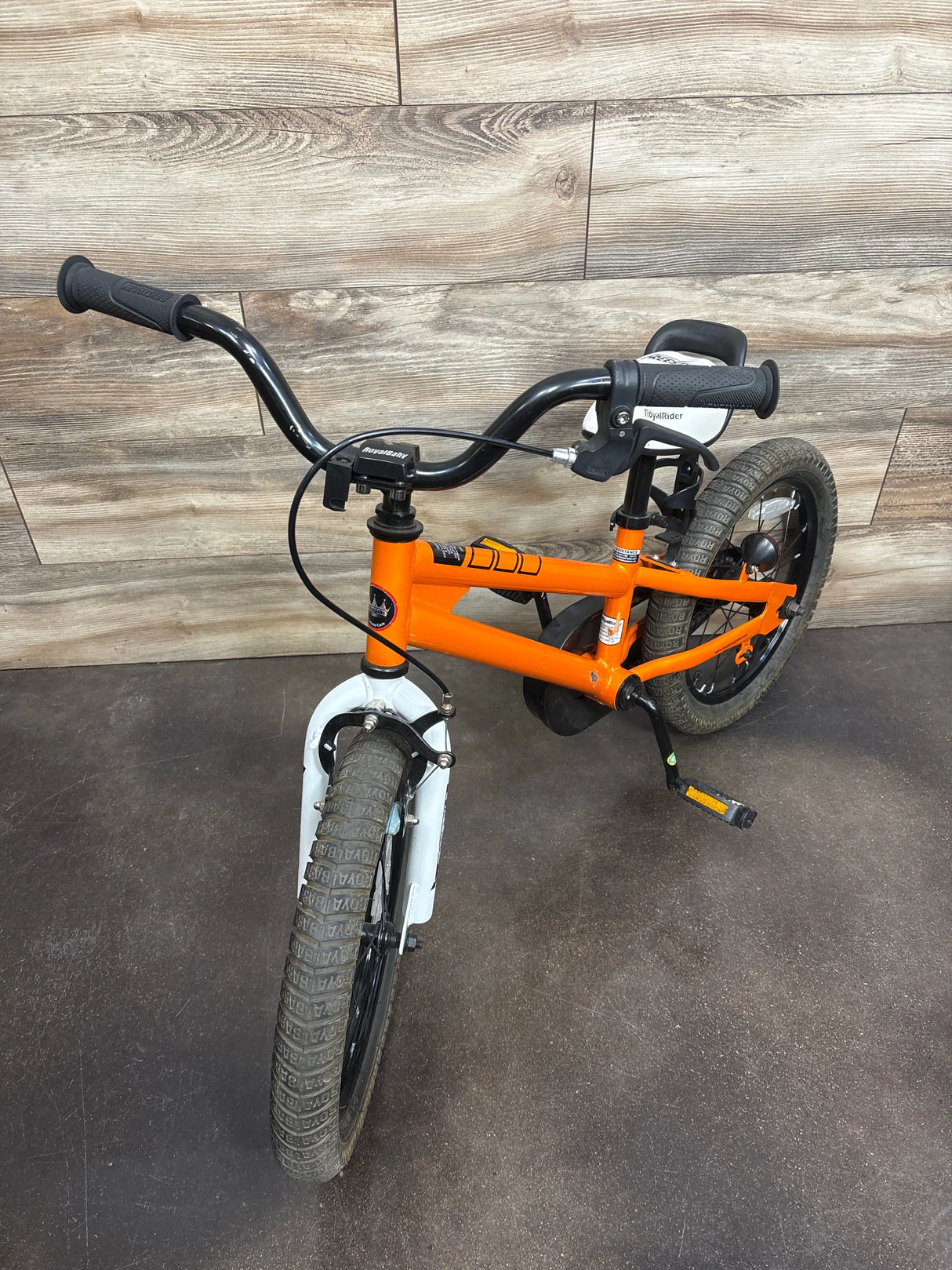 Orange RoyalBaby kids bike with training wheels, black handlebars, and white fenders on wood floor