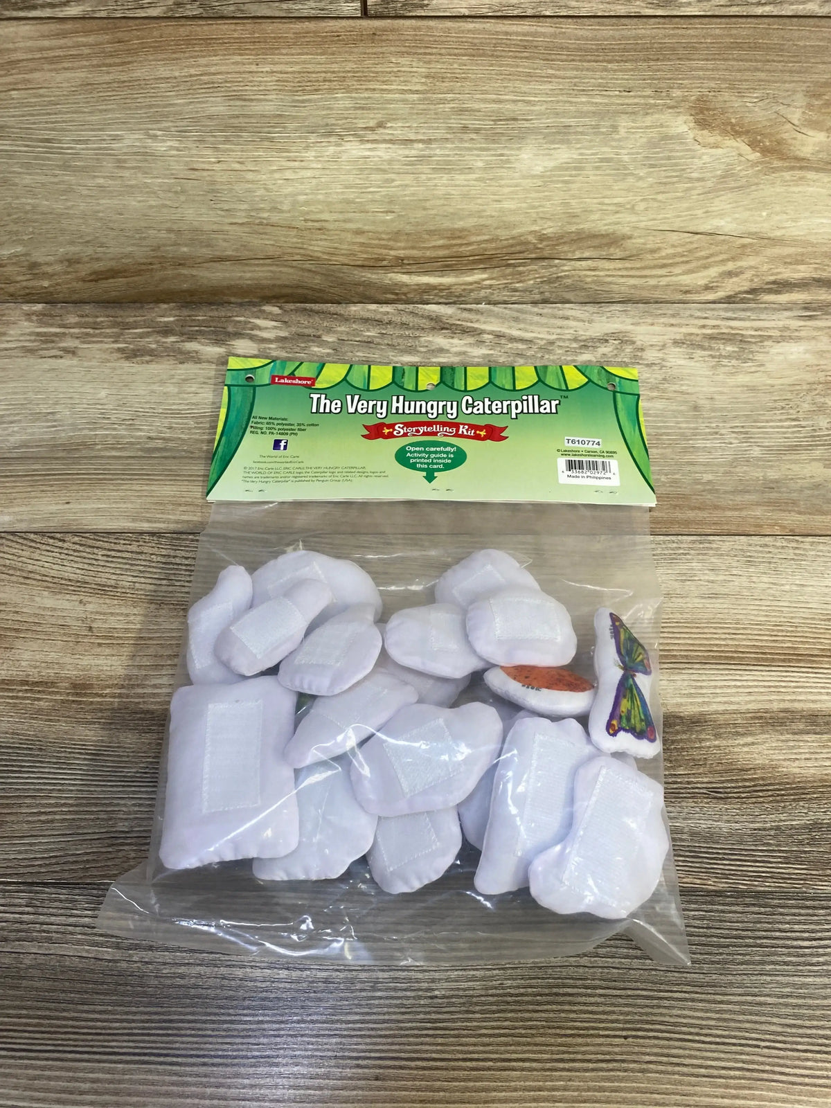 Top-down view of The Very Hungry Caterpillar storytelling kit showing Velcro-backed white felt pieces and printed food and butterfly in a clear bag on a wood tabletop