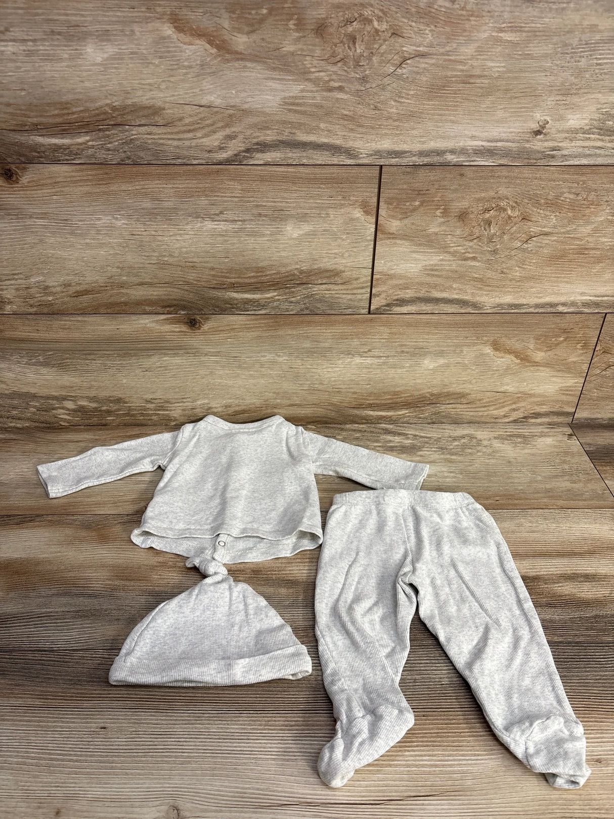 Flat lay of gray ribbed 3-piece baby set showing long-sleeve top with knotted hem, footed pants, and knotted hat on wood floor