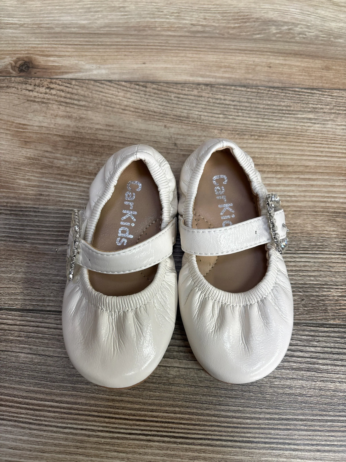 White baby Mary Jane shoes with elastic edges and decorative rhinestone buckles on wooden floor