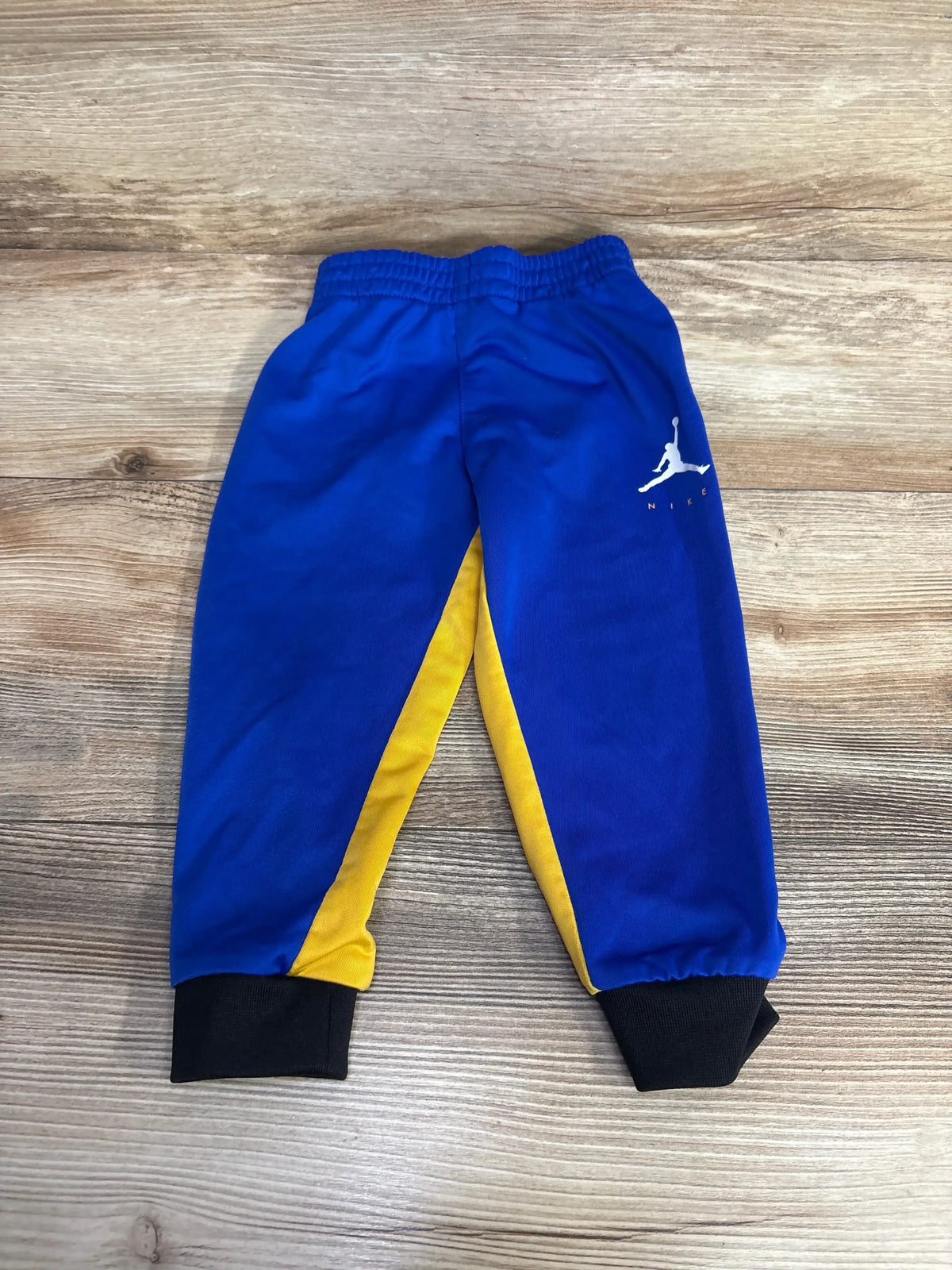 Flat lay of 2T Jordan pull-on pants showing blue fabric, yellow side panels, black ankle cuffs, and white Jumpman logo on wood floor