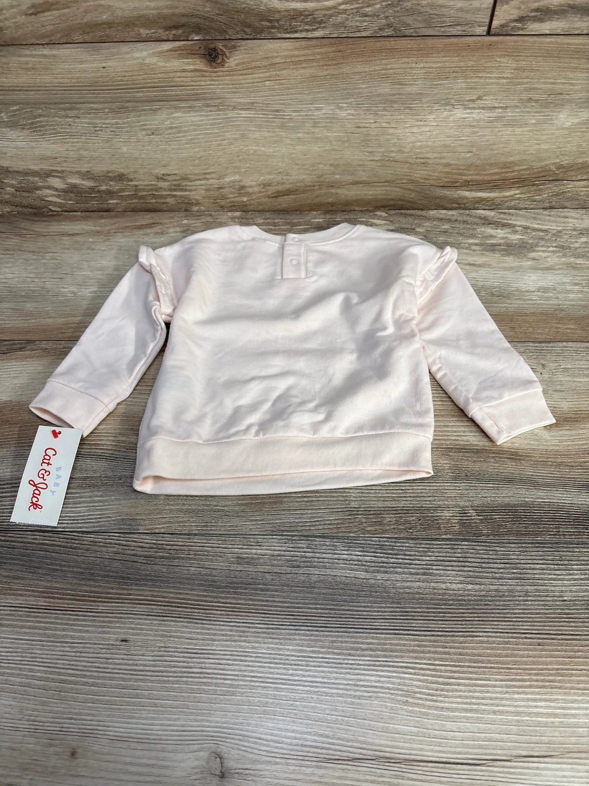 Back view of pink Cat & Jack baby sweatshirt showing ruffle shoulders and snap closure at neck on wood-plank background
