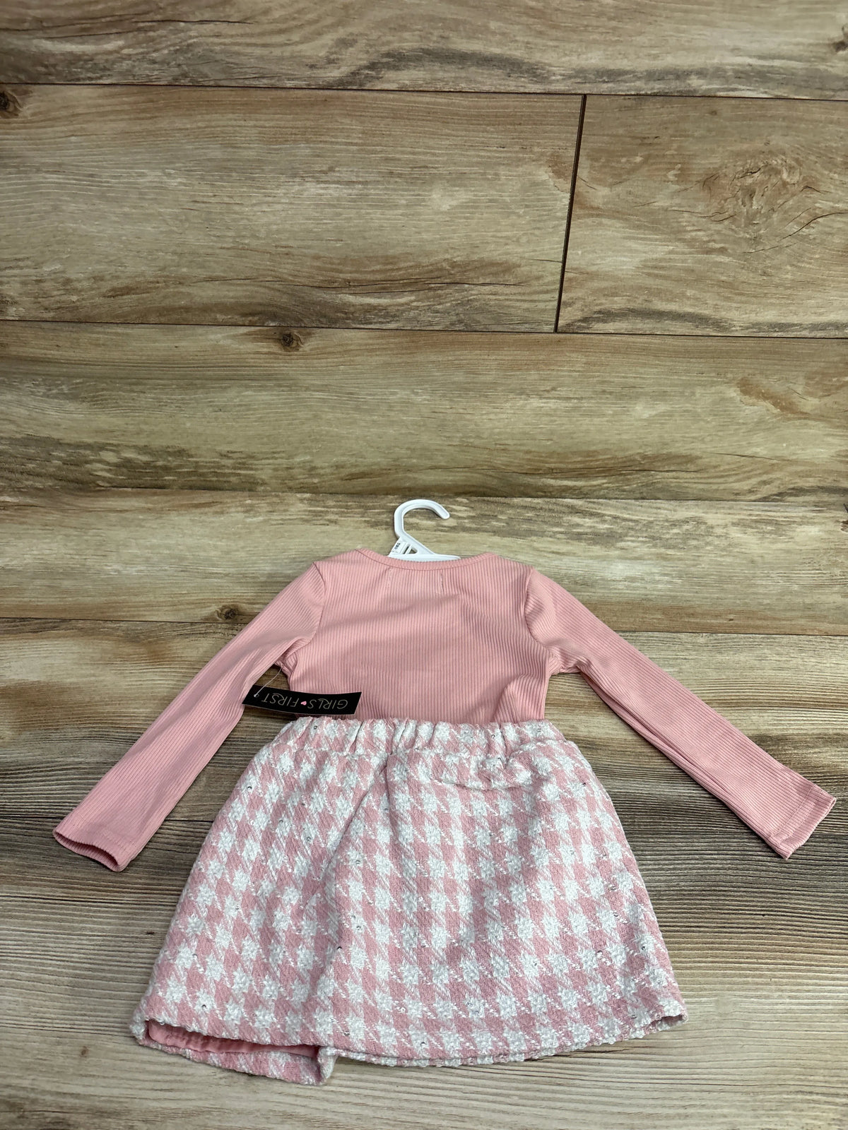 Back view of pink long sleeve tartan skirt dress showcasing patterned skirt on a hanger against a wood floor