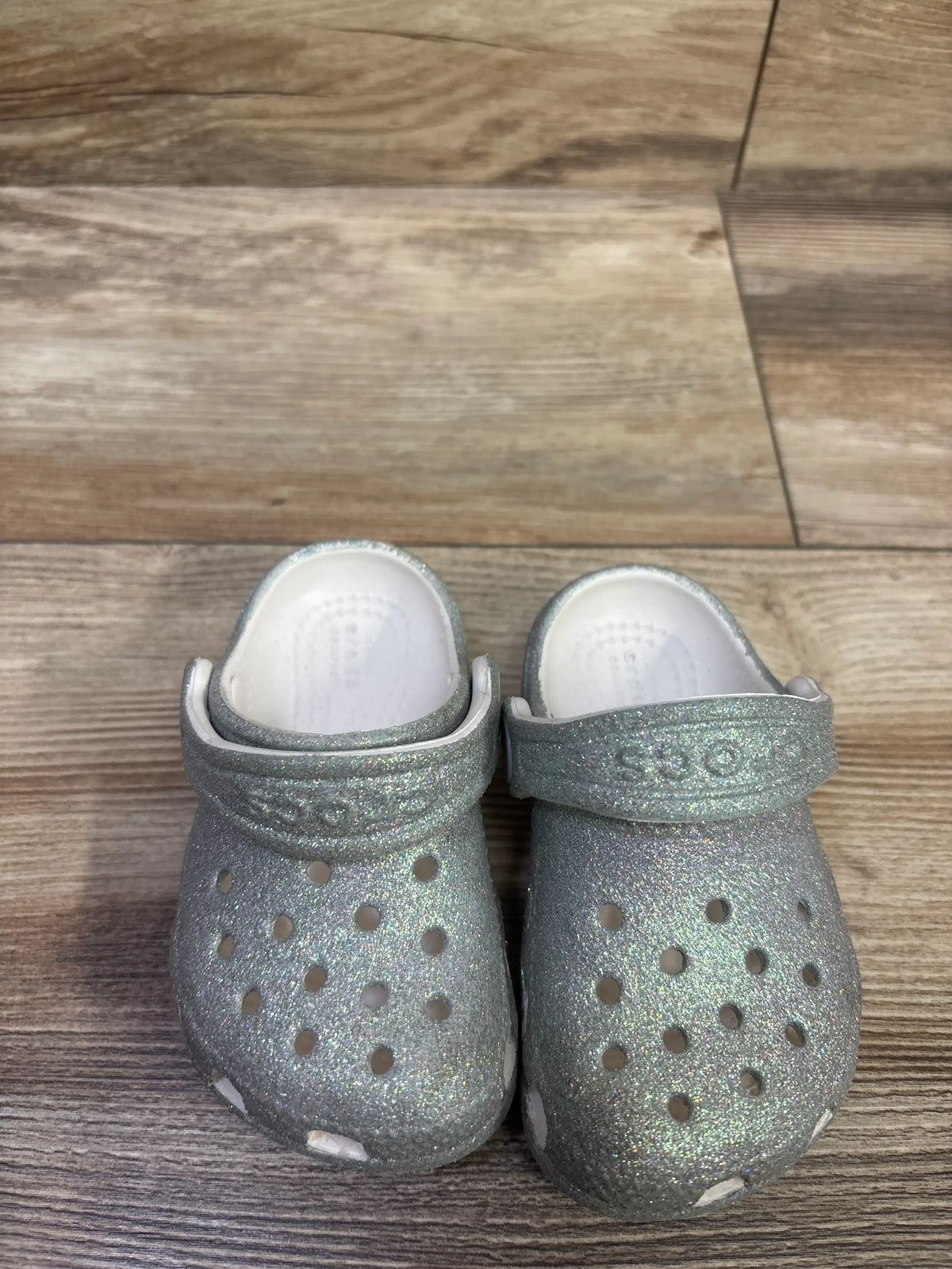 Front view of silver glitter Crocs Classic clogs showcasing ventilation holes and heel straps on a wood floor