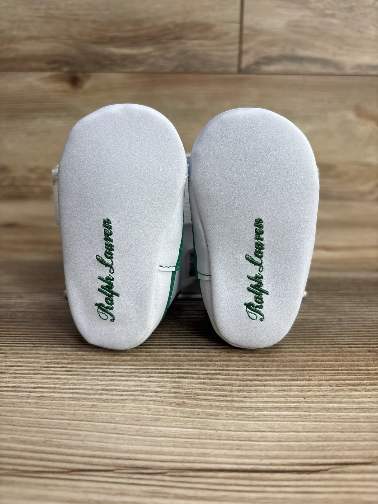 Pair of white Ralph Lauren baby shoes with green logo on soles on wooden background