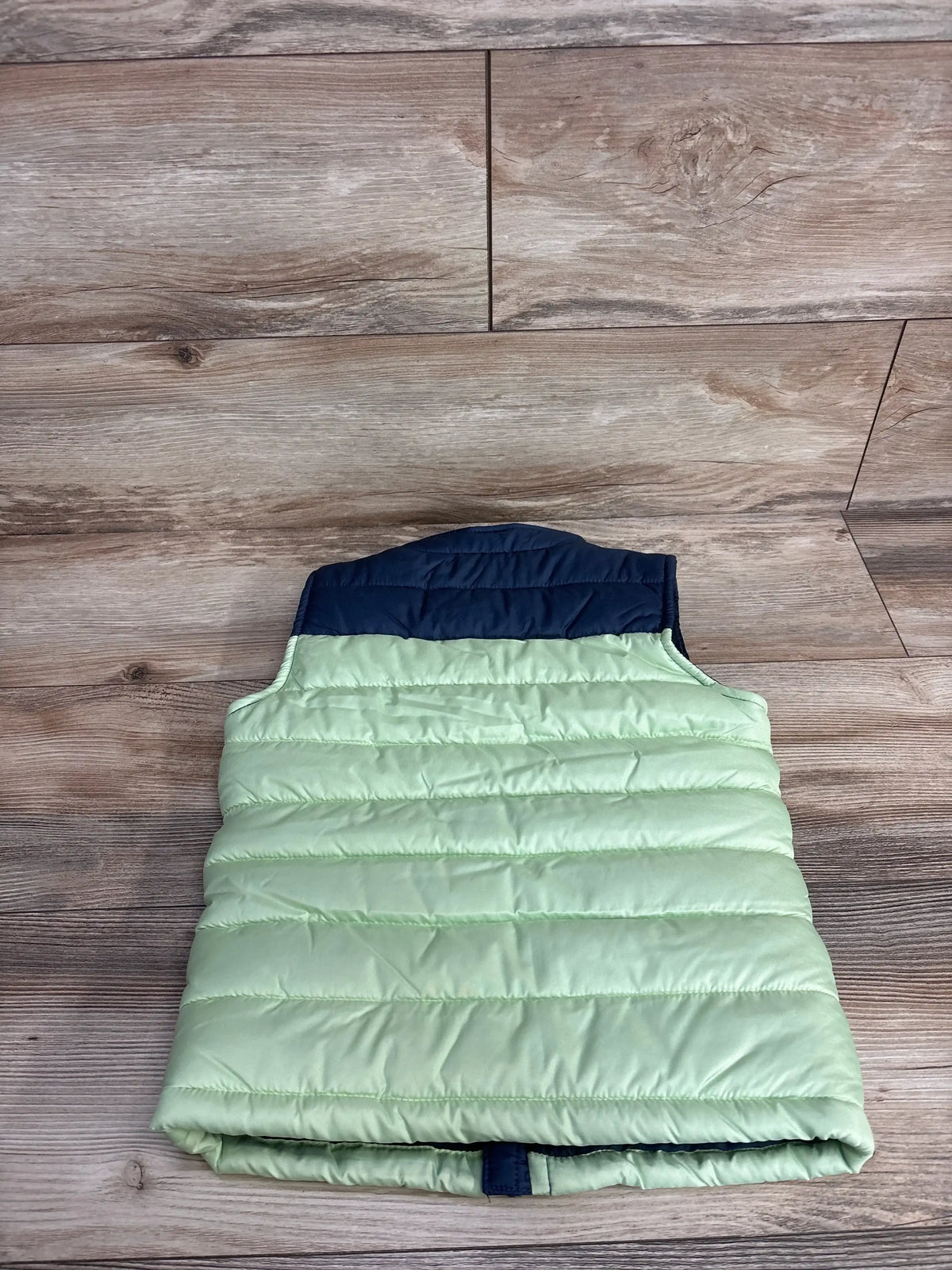 Light green kids puffer vest with navy collar, laid flat on wood background