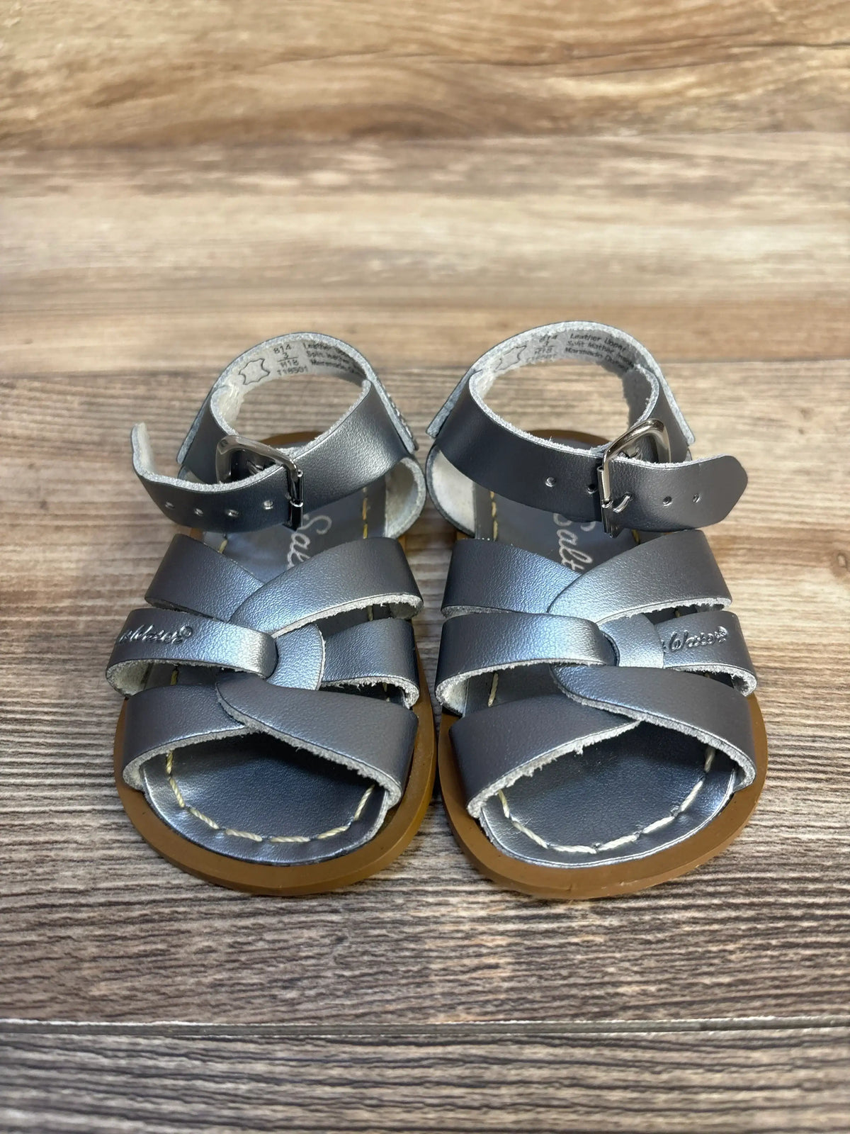 Front view of Salt-Water Original toddler sandals, silver size 3C, crisscross leather straps and ankle buckles on a wood floor