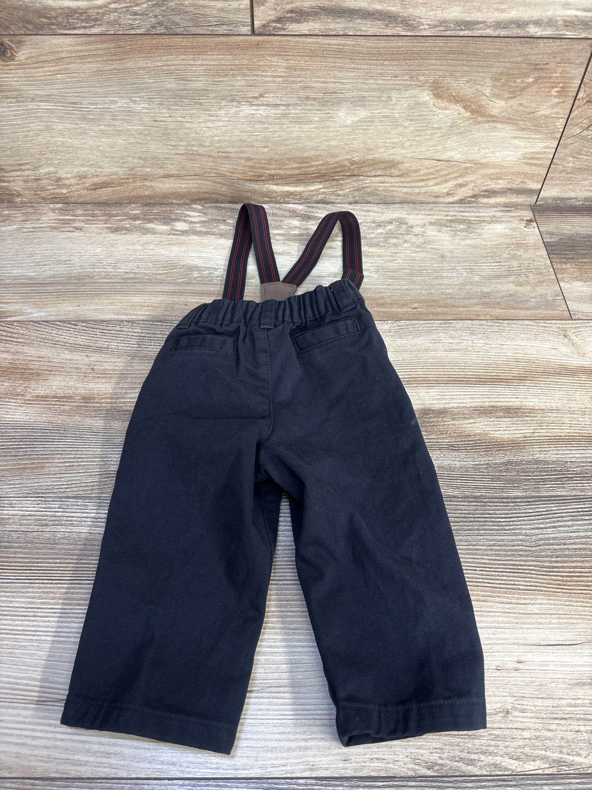 Back view of black suspender pants, 9M, with elastic waist, faux back pockets and red-striped suspenders on light wood floor