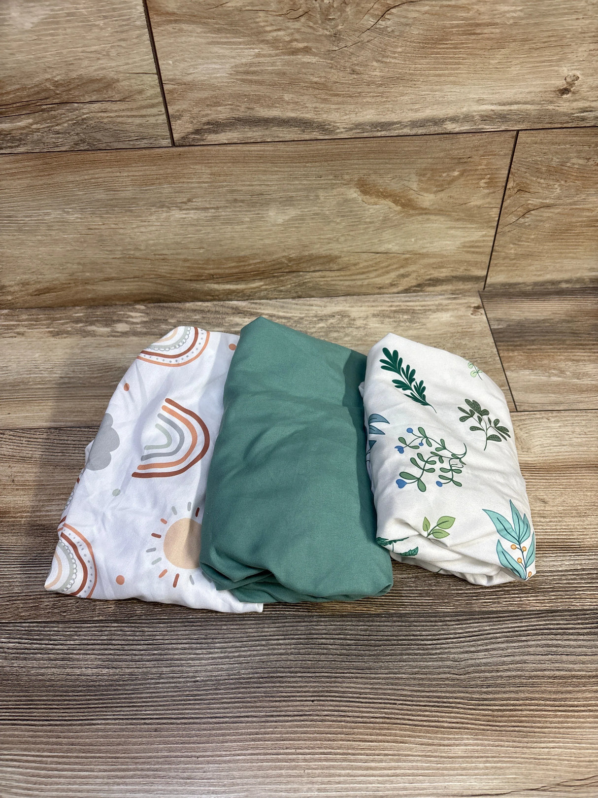 Folded view of Haleeone 3-pack fitted bassinet sheets showcasing solid green and two printed (rainbow and leaf) designs on a wood background