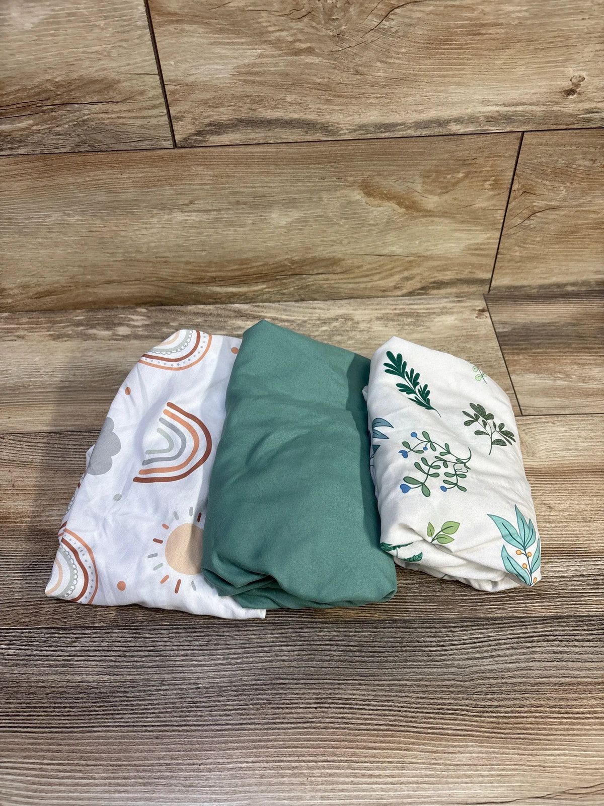 Folded view of Haleeone 3-pack fitted bassinet sheets showcasing solid green and two printed (rainbow and leaf) designs on a wood background