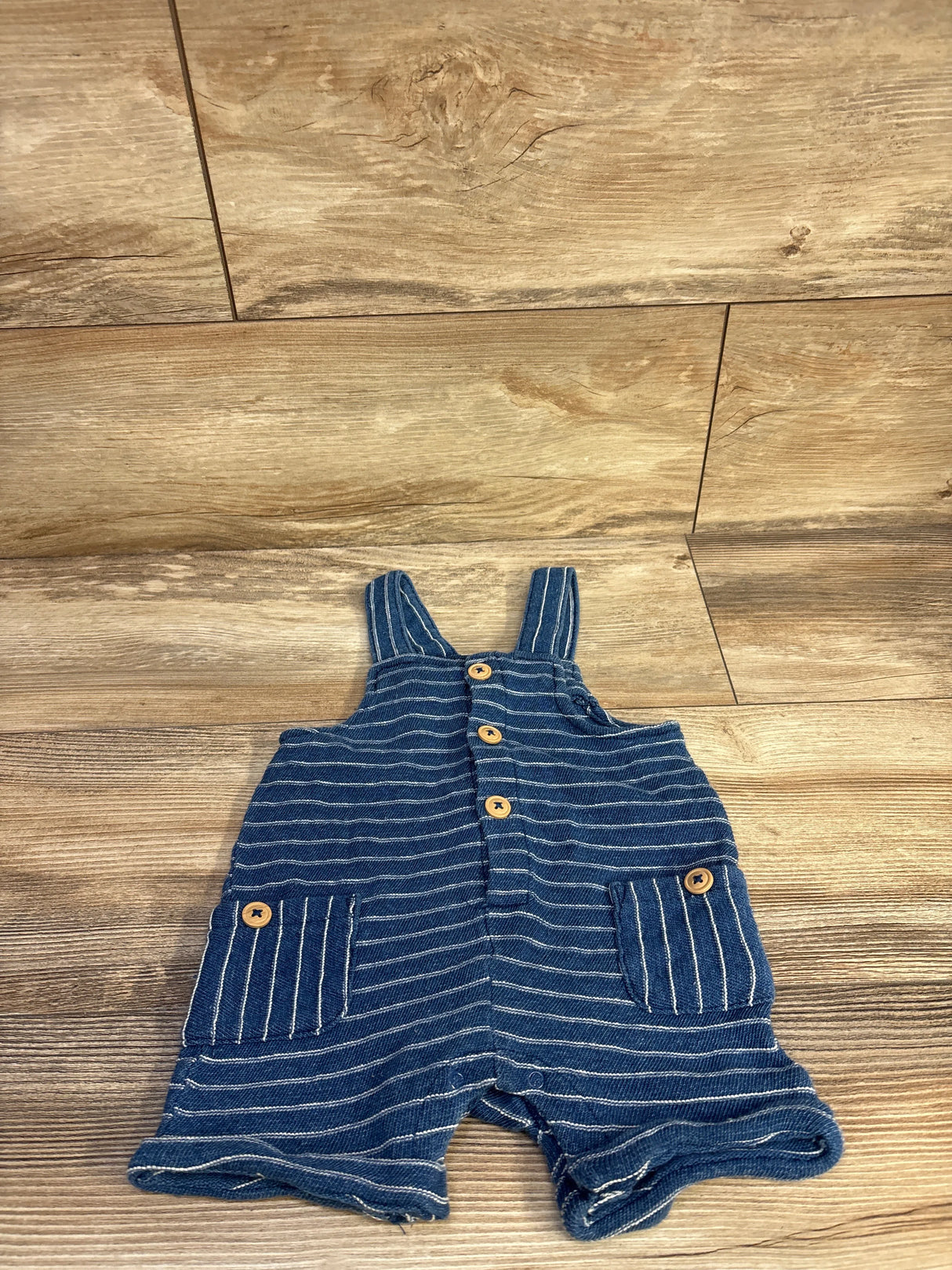 Flat lay of blue Cat & Jack striped henley shortalls showcasing front button placket and two patch pockets with wooden-look buttons on wood floor background