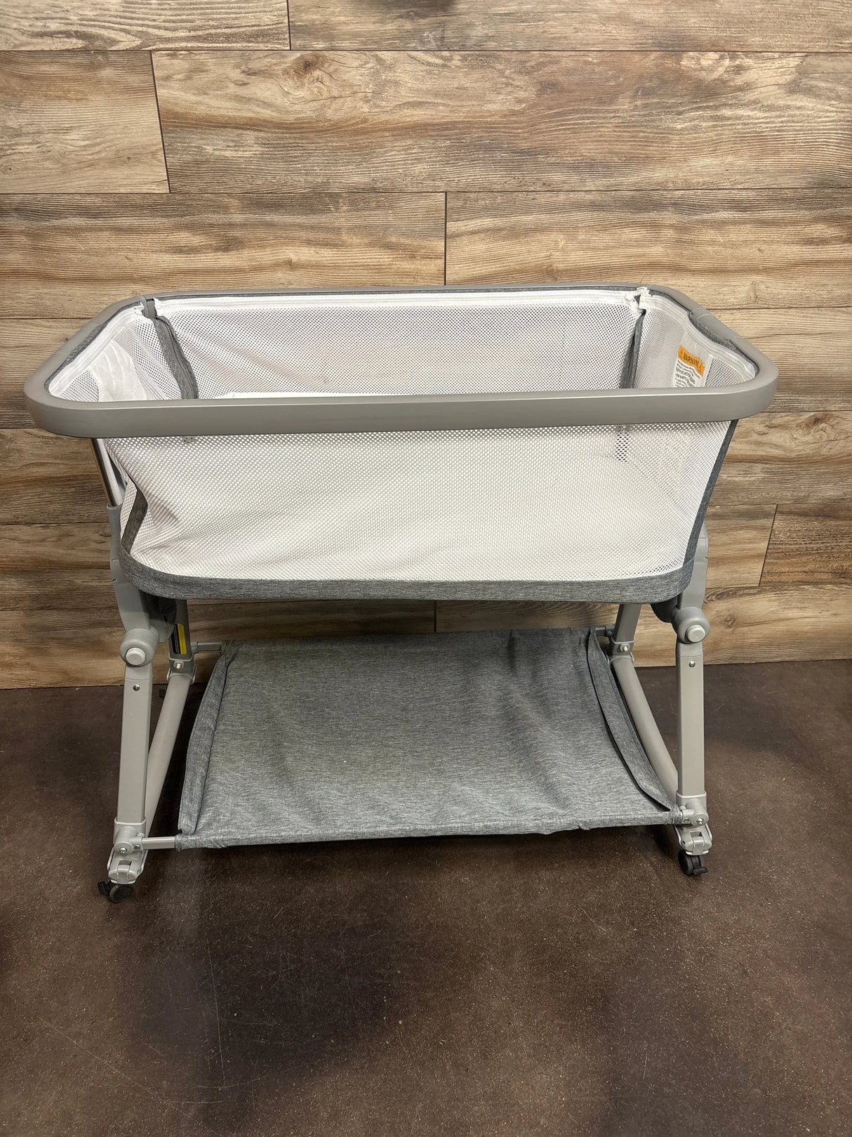Front view of Lil Pengyu Bedtime Bliss Bassinet in grey mesh with grey storage shelf and wheels in front of dark wood plank wall