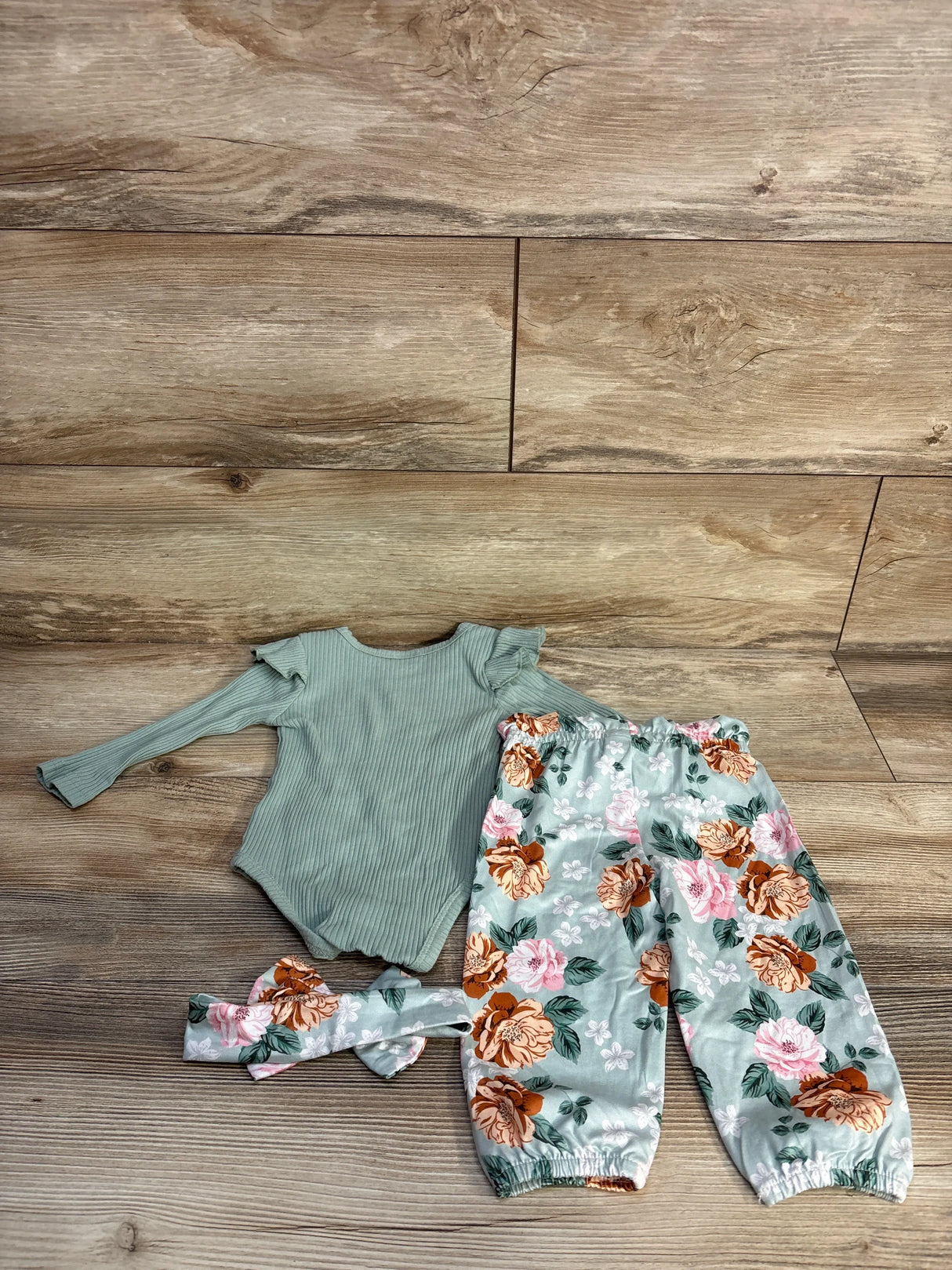 Flat lay of 4pc baby set: green ribbed long-sleeve bodysuit, mint floral elastic-waist pants, matching headband on wood floor