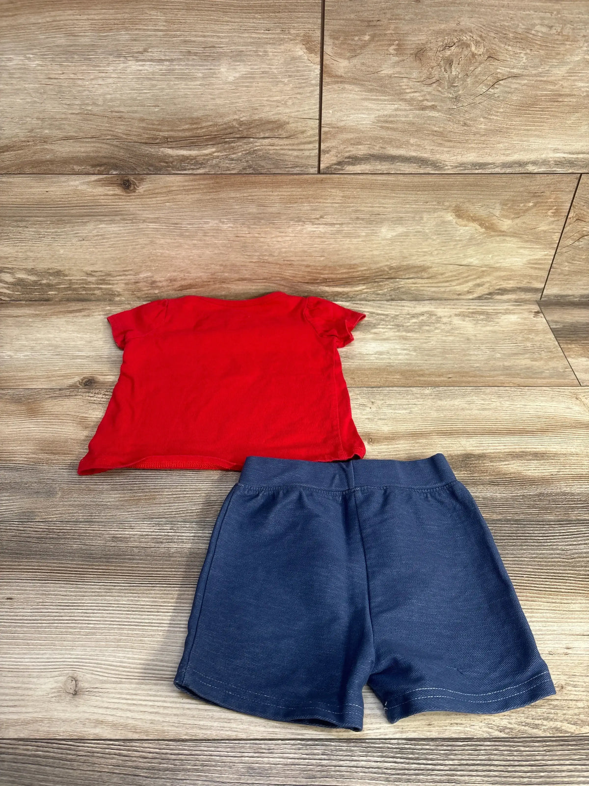 Flat lay of toddler 2-piece shirt and shorts set showcasing red short-sleeve top and navy elastic-waist shorts on wood background