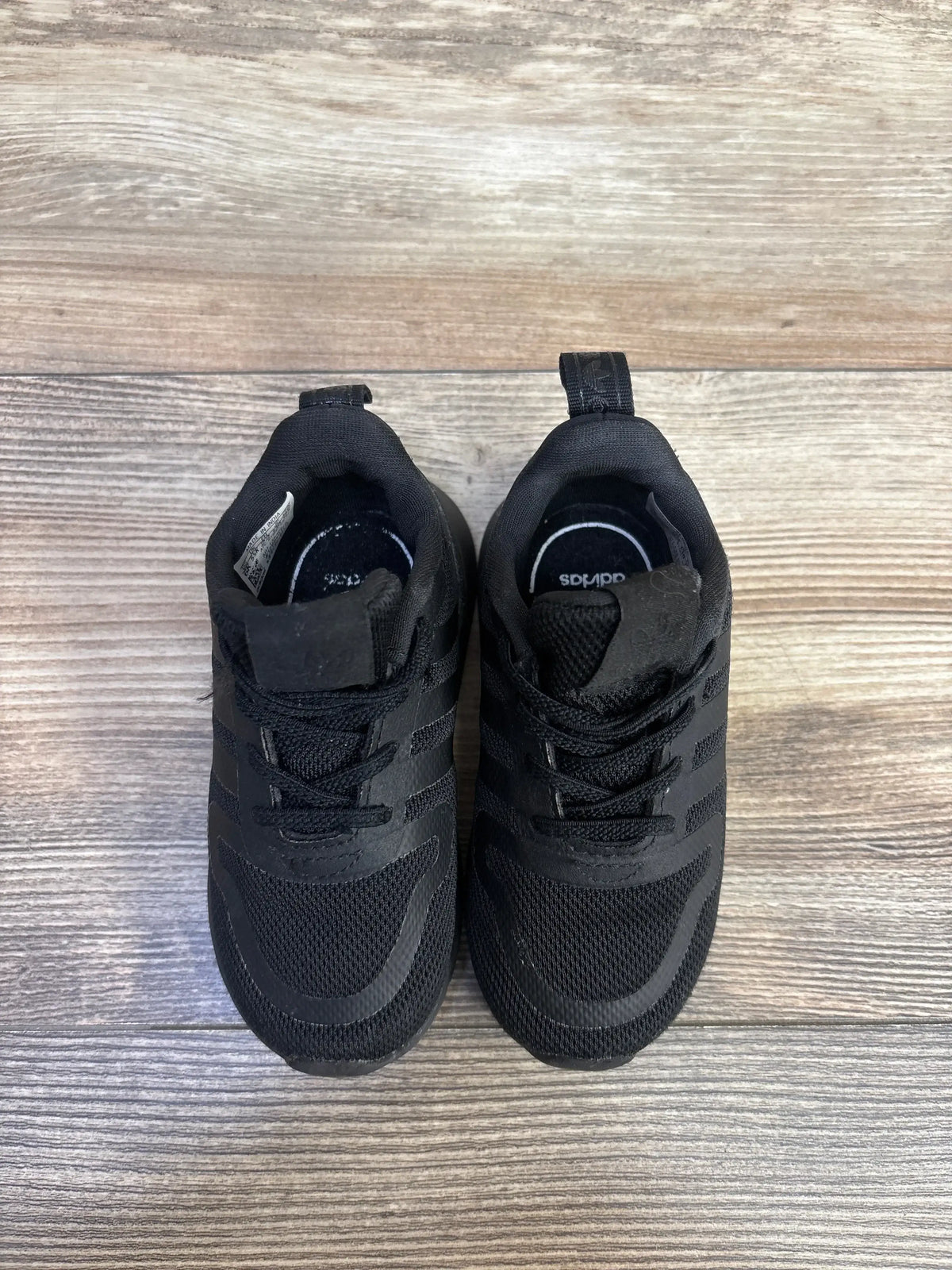 Top-down view of Adidas Multix I toddler black sneakers showing mesh upper, three stripes, and laces on light wood floor