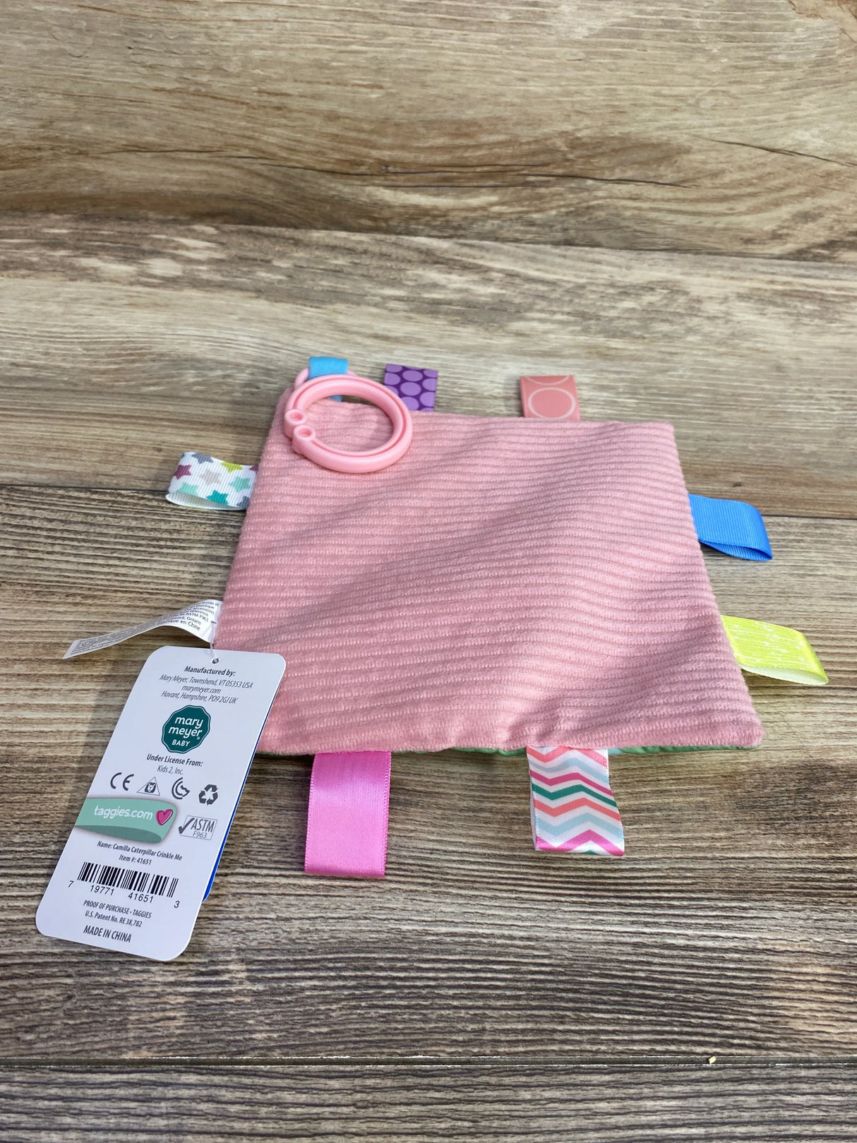 Top-down view of Taggies Camilla Caterpillar Crinkle Me baby blanket showing pink ribbed fabric, colorful ribbon tags, and attached pink teething ring on wood background