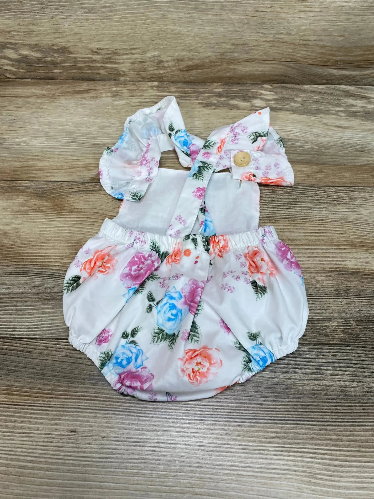 Top view of white floral bubble romper (3-6m) with ruffle shoulder straps, wooden button and elastic legs on wood floor