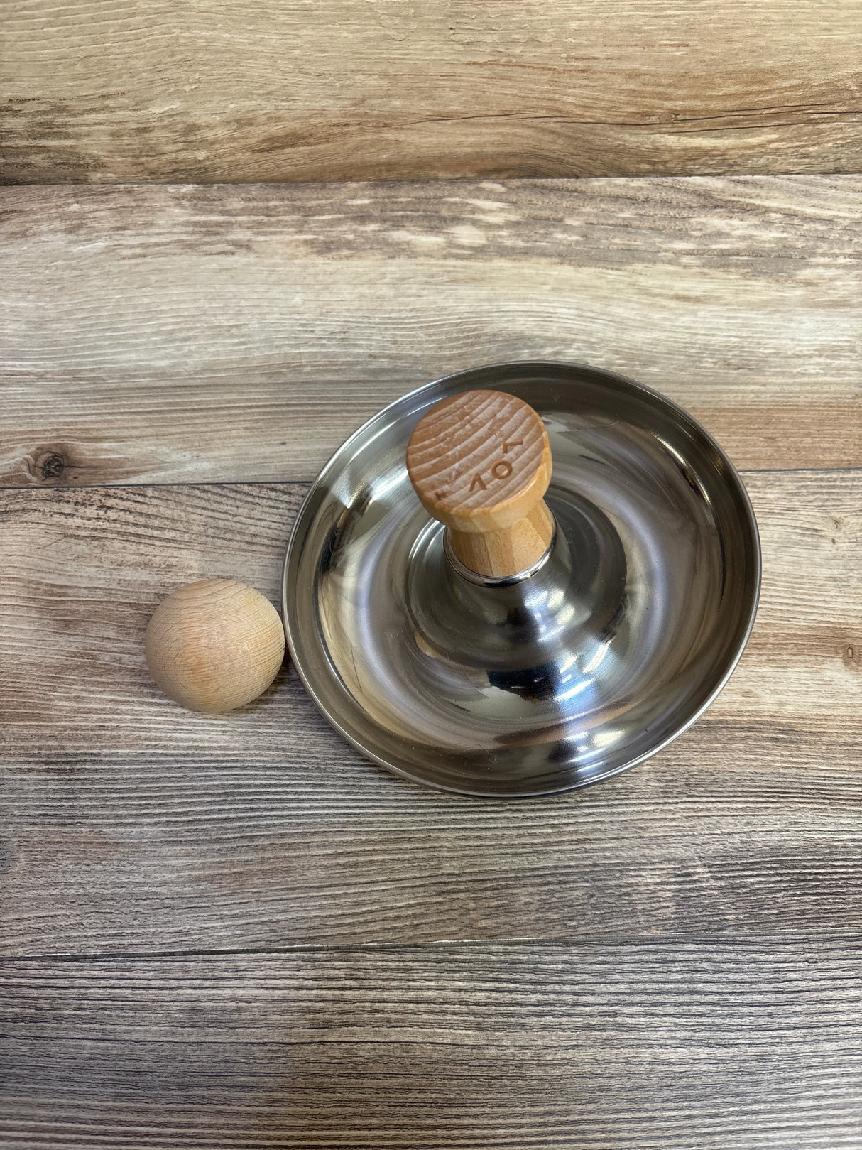 Angled view of Lovevery Stainless Steel Tip and Turn showcasing a stainless steel base, wooden handle, and ball on wood surface