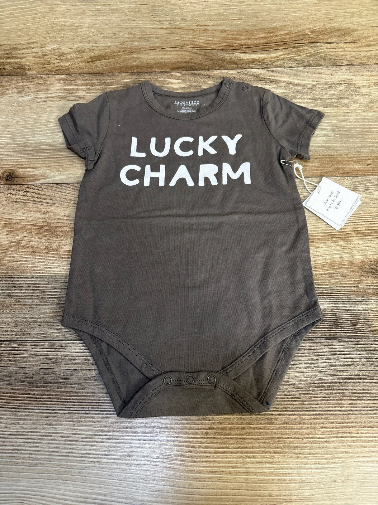 Flat lay of Face To Face Lucky Charm bodysuit in grey with white text print and hangtag attached on wood plank floor