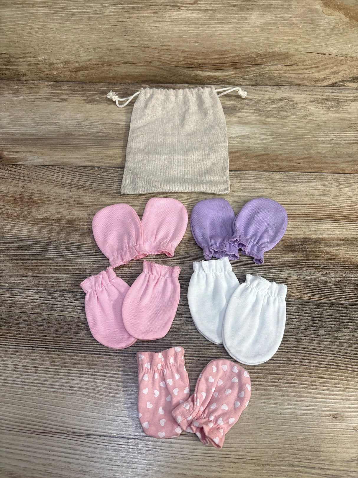 Flat lay of 5 infant mitten pairs in pink, purple and white; one pink heart-print; elastic cuffs with beige pouch on wood