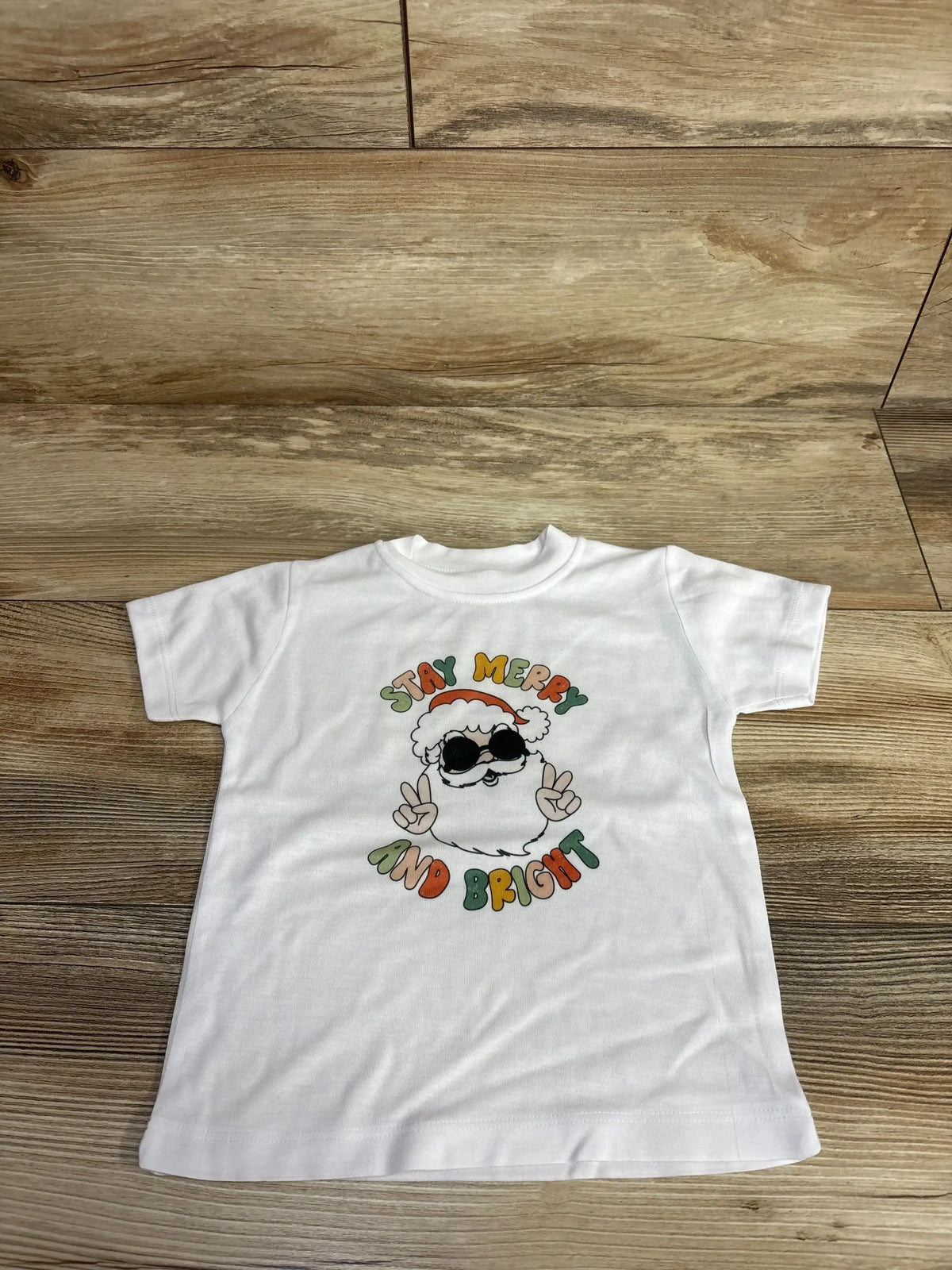 White kids Christmas tee with Santa graphic saying 'Stay Merry and Bright', laid on wood floor