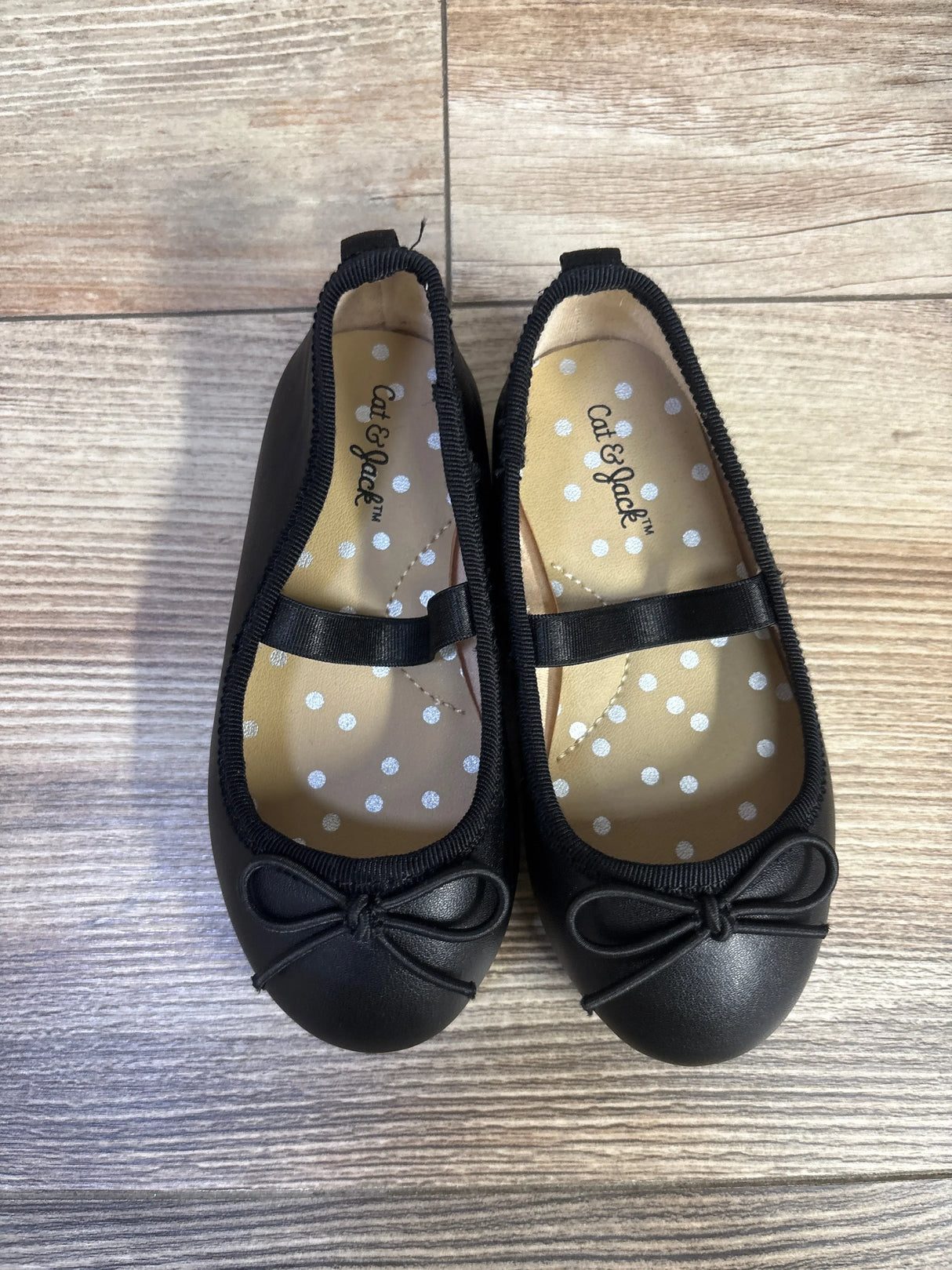 Top-down view of Cat & Jack Nora ballet flats in black with front bow and elastic strap, polka-dot insole on wood floor