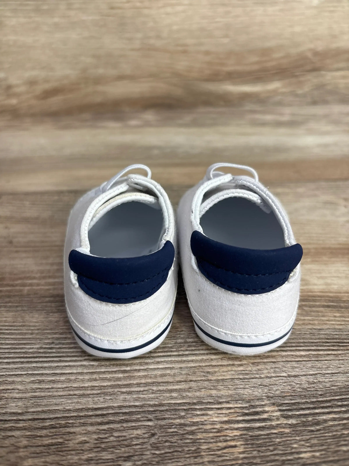 Back view of baby white canvas shoes with navy blue heel accents on wooden surface