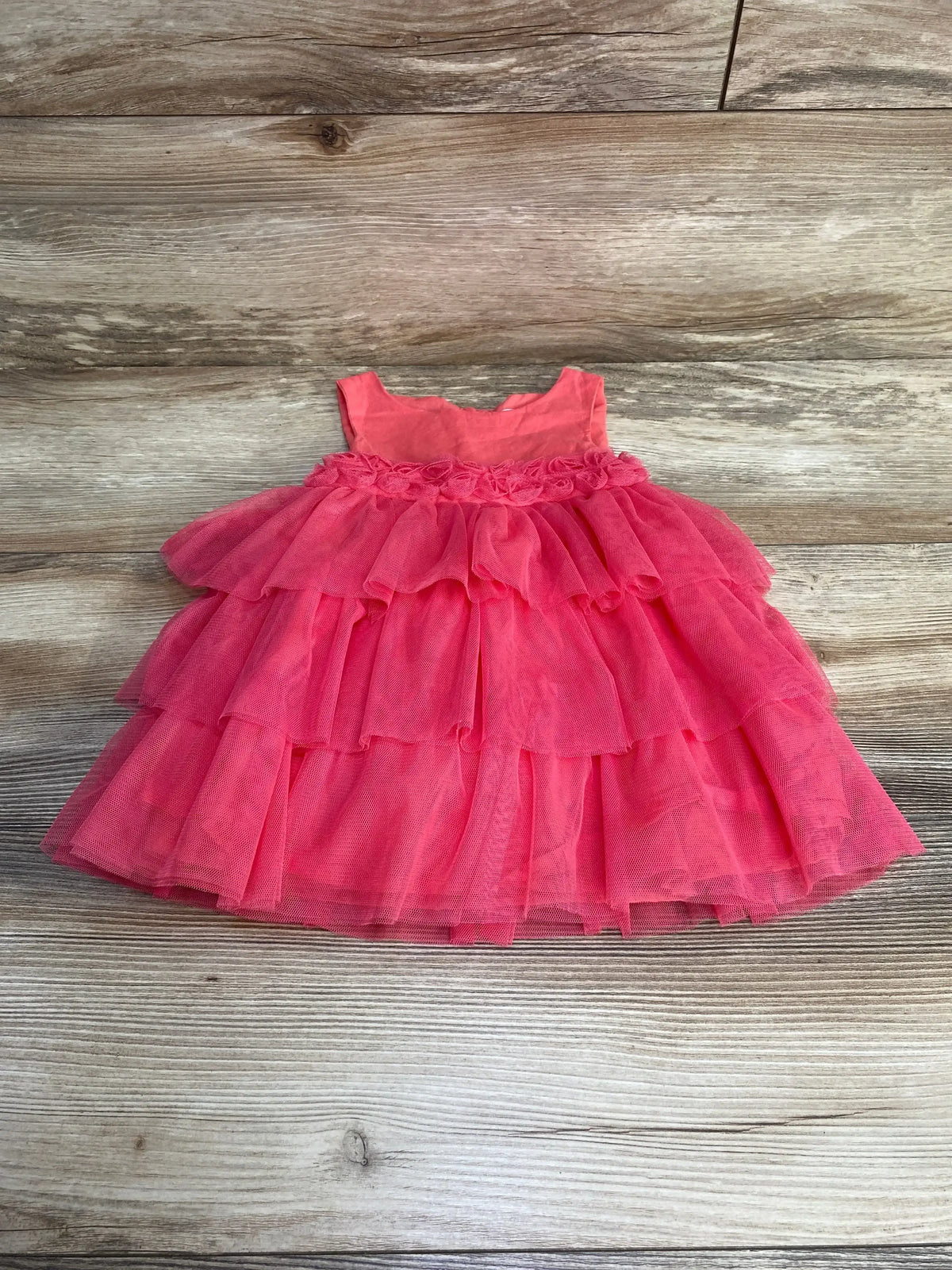 Flat lay of pink tulle baby dress showcasing three-tier ruffled skirt and rosette waist trim on wood background