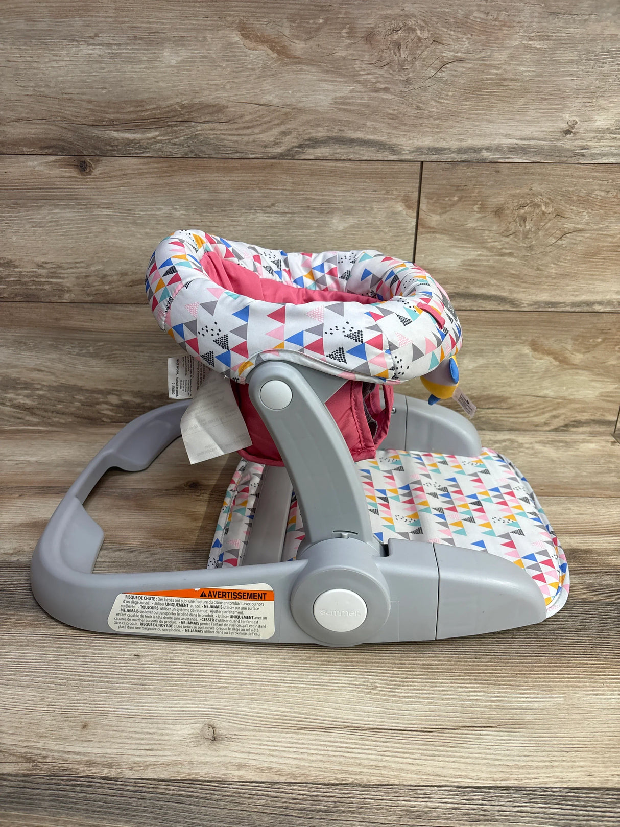 Pink and multicolor triangle patterned baby floor seat with gray plastic frame on wooden surface