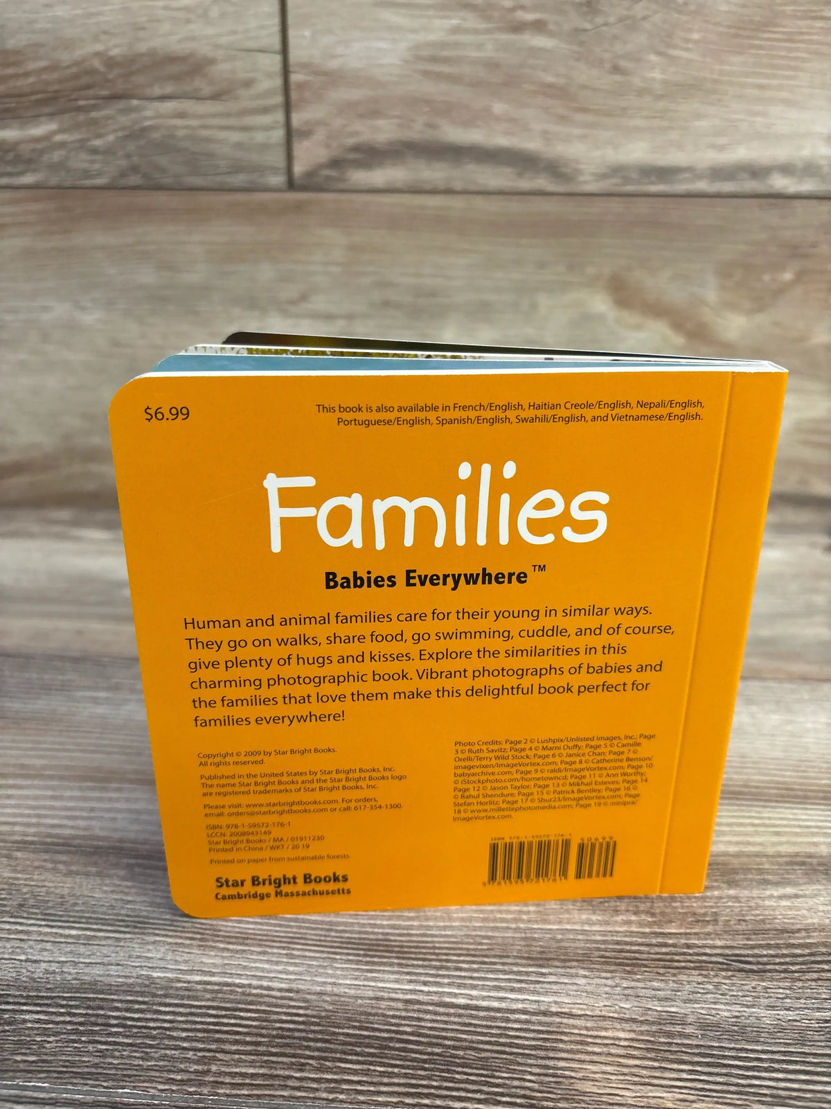 Back view of Families (Babies Everywhere) board book showing orange back cover with title, blurb, and barcode on wood table