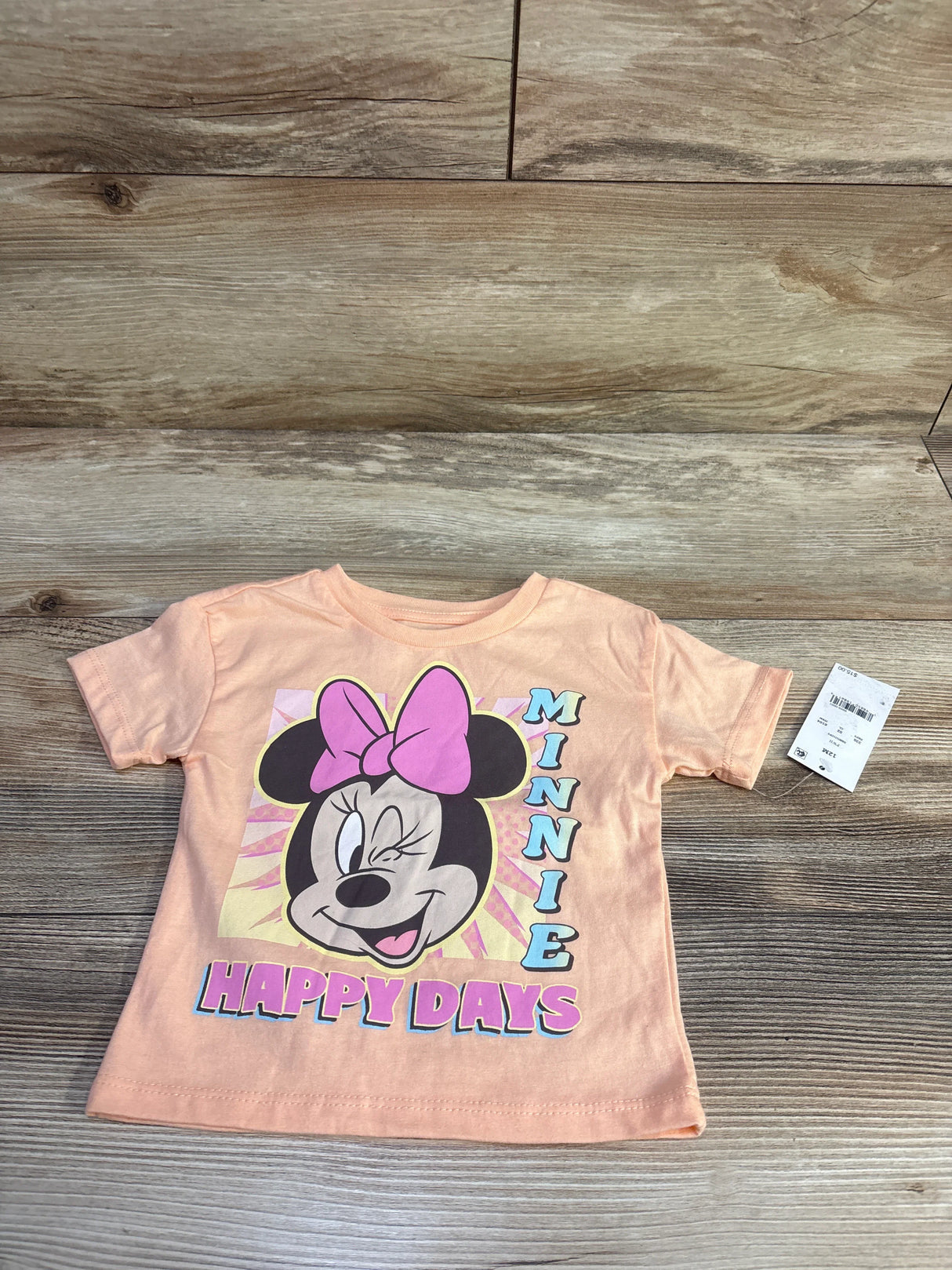 Flat lay of peach Minnie Mouse toddler shirt with winking Minnie face, pink bow, shows MINNIE and HAPPY DAYS on wood background
